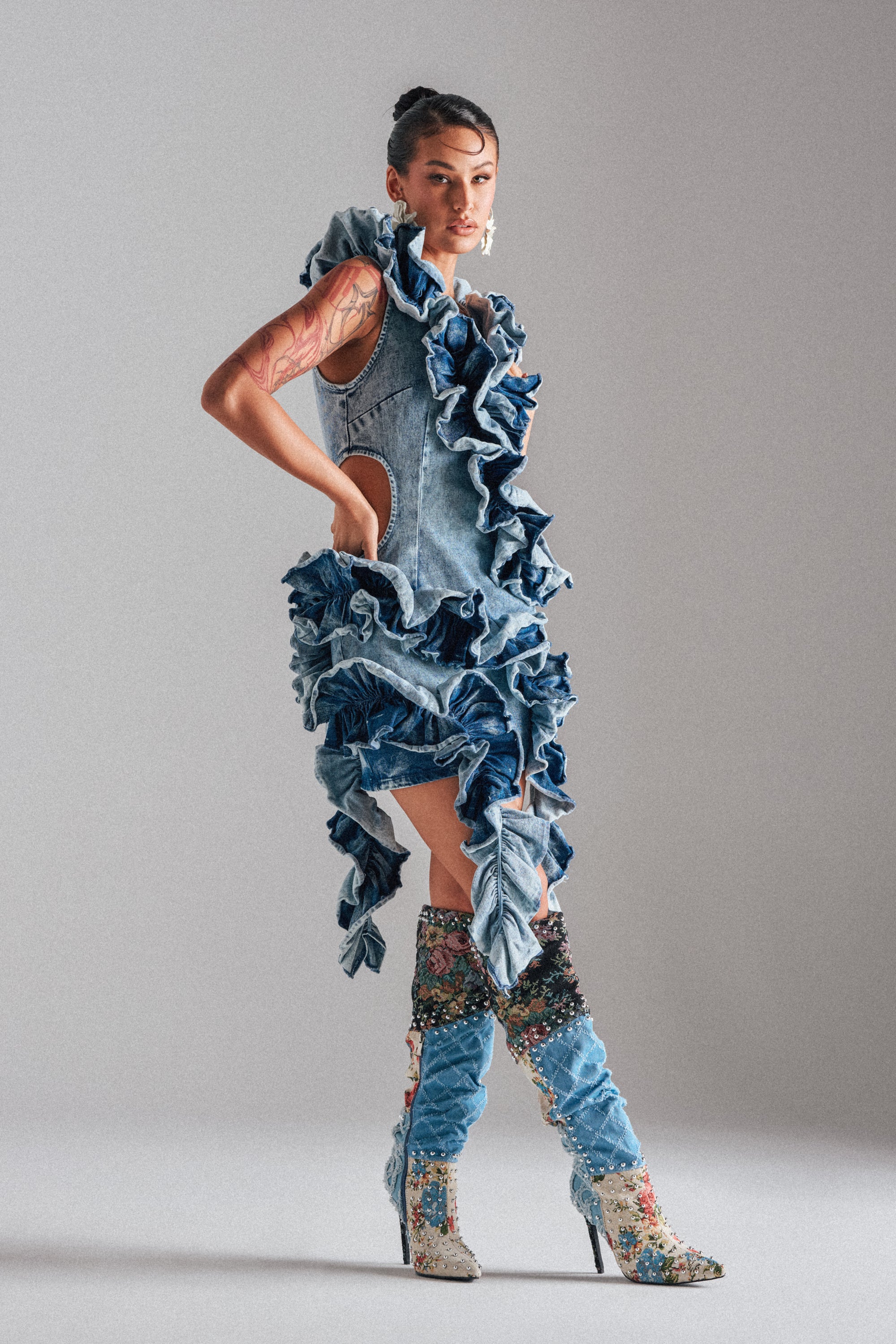 A tattooed model poses in the SAY IT AGAIN RUFFLED DENIM MINI DRESS, a sleeveless statement piece with ruffles, styled with tall embellished heeled boots. The plain light background showcases the dress's texture and bold silhouette. 2