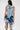 A woman with long dark hair is seen from the back wearing the SAY IT AGAIN RUFFLED DENIM MINI DRESS with cutouts. She holds a small white handbag and stands against a plain light background.