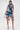 A woman faces forward in the SAY IT AGAIN RUFFLED DENIM MINI DRESS, featuring dramatic ruffles and a high-low hem. She pairs it with a silver handbag and pointed blue heels, standing against a plain white background.