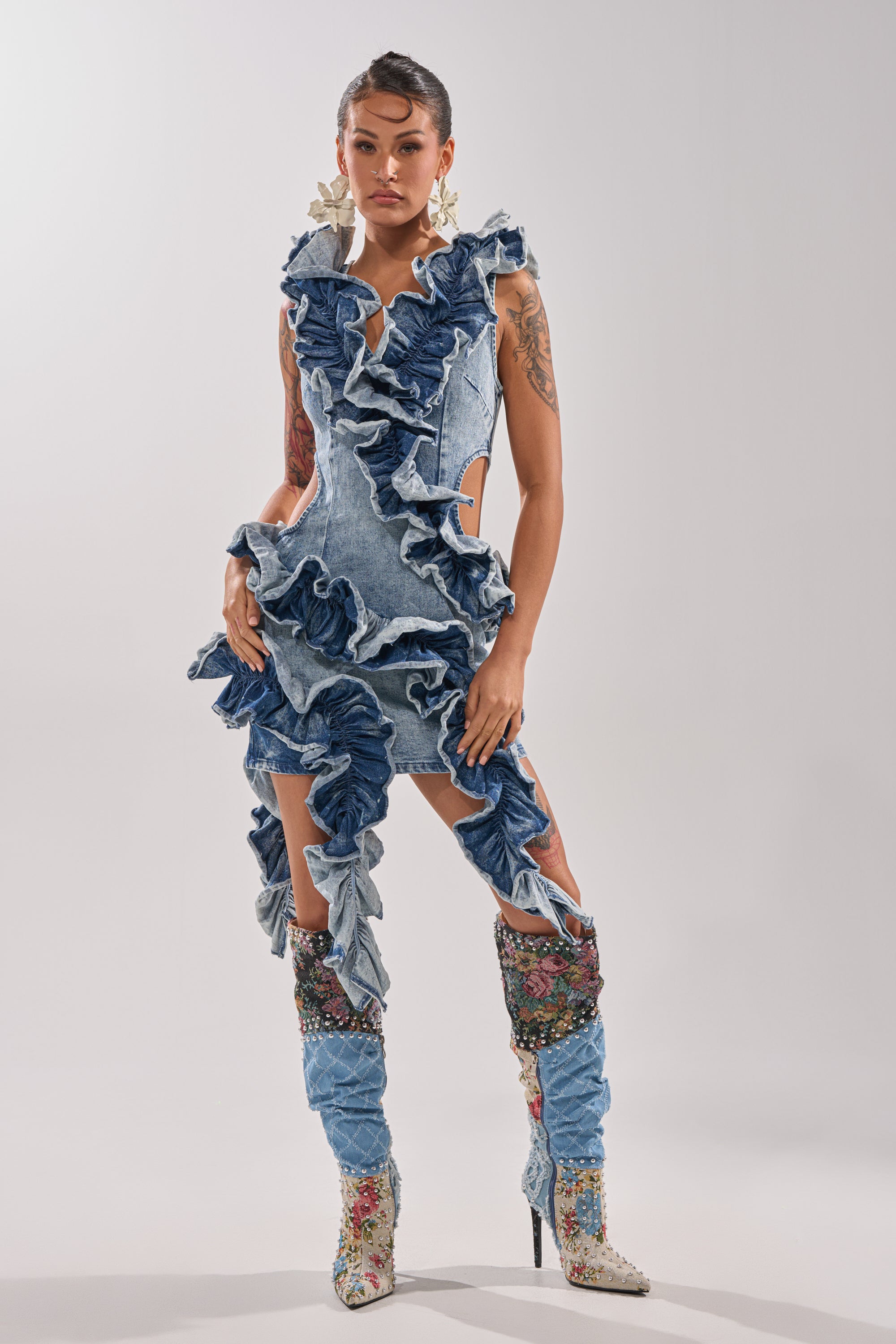 A woman stands confidently in the SAY IT AGAIN RUFFLED DENIM MINI DRESS, featuring dramatic blue frills and high slits, paired with knee-high embroidered boots and large earrings against a plain light background. 8
