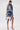 A woman stands on a white background wearing the SAY IT AGAIN RUFFLED DENIM MINI DRESS featuring dramatic ruffles, side cutouts, and asymmetrical layers. She holds a small white handbag and pairs the look with blue high heels.
