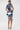 A woman wears the SAY IT AGAIN RUFFLED DENIM MINI DRESS, featuring dramatic frill details, paired with a small silver handbag and light blue high heels against a plain white background.
