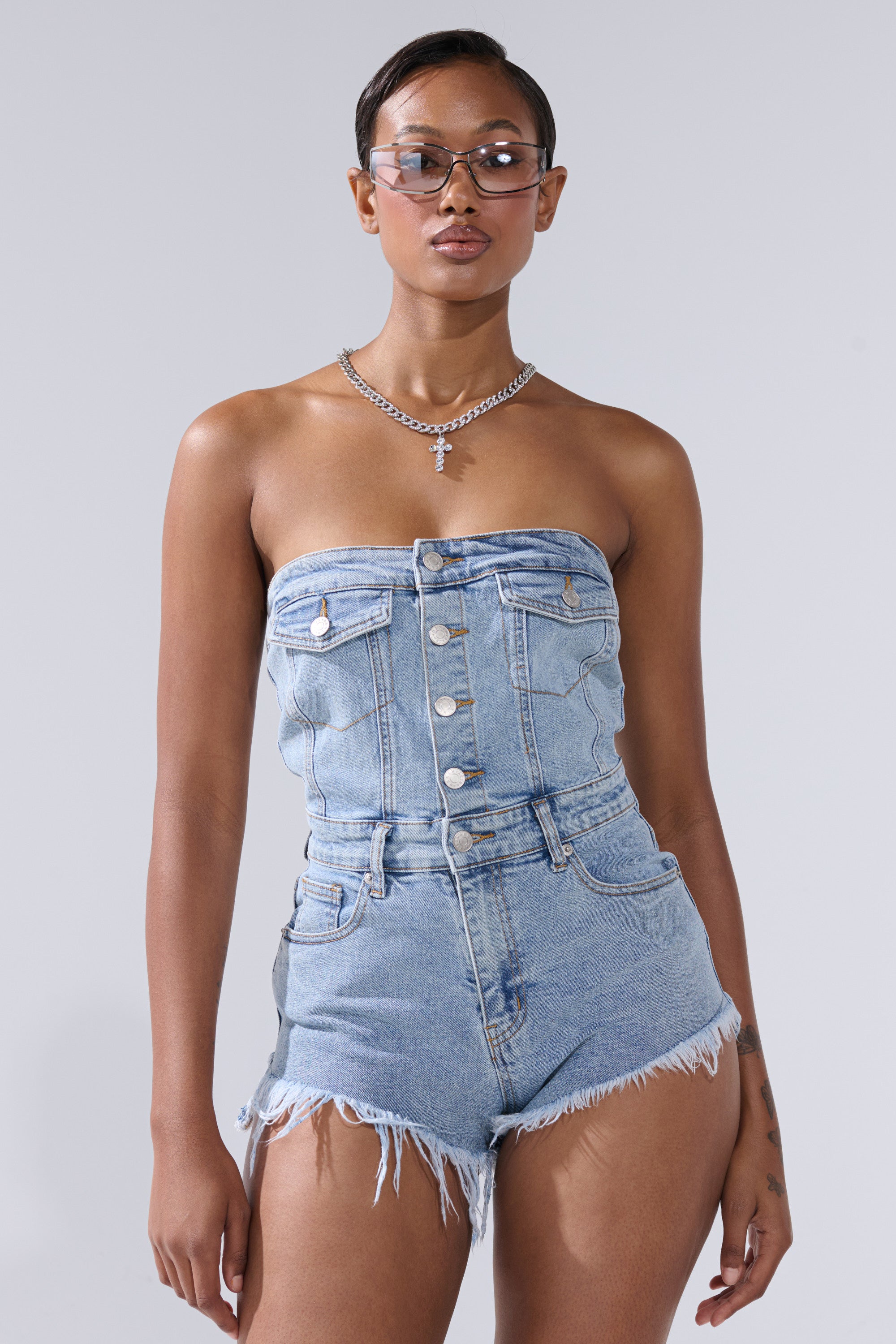 A woman models the COOL IT COWBOY DENIM ROMPER, featuring a strapless top and high-waisted frayed shorts, styled with a silver chain necklace, cross pendant, and clear-framed glasses against a plain gray background.