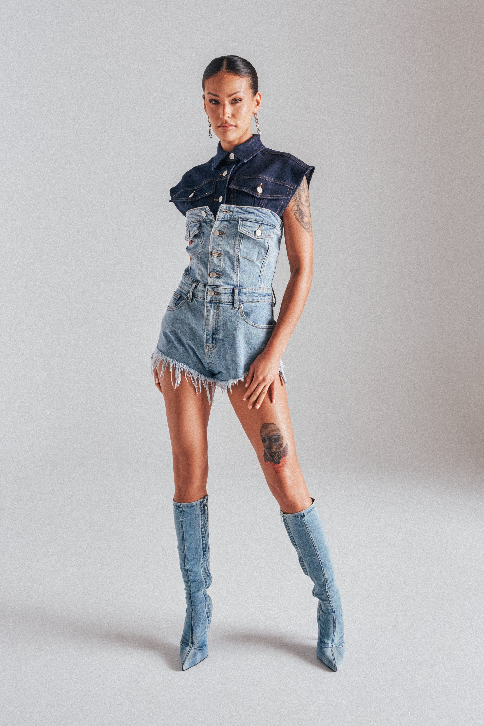 A woman poses confidently against a plain background in the COOL IT COWBOY DENIM ROMPER with frayed edges, styled with matching knee-high denim boots, hoop earrings, and showcasing tattoos on her left arm and thigh. 1