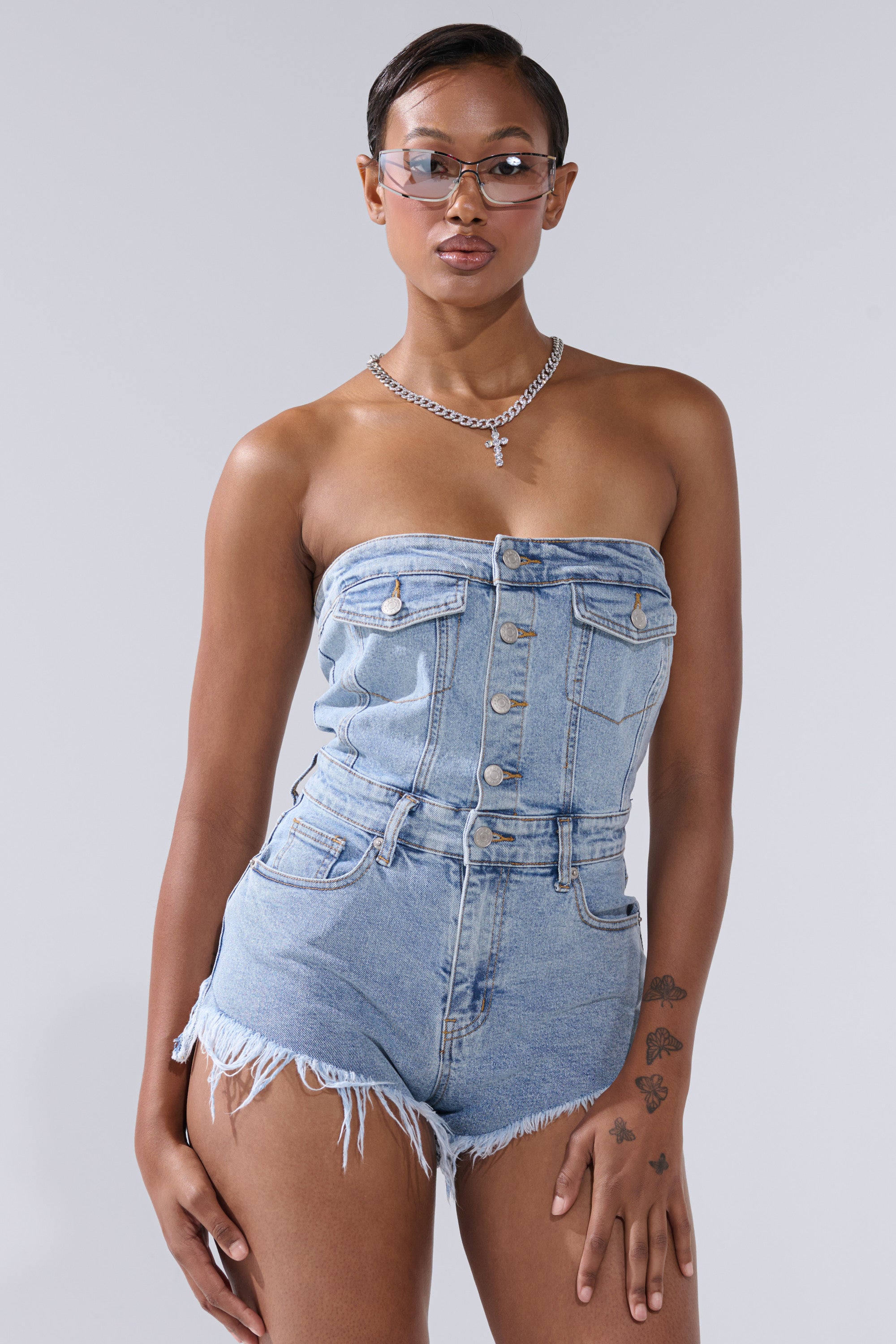 A woman with short hair, a hand tattoo, silver chain necklace, and clear sunglasses poses confidently in the COOL IT COWBOY DENIM ROMPER—strapless with frayed shorts—giving this western piece a fresh, modern edge against a light gray backdrop.