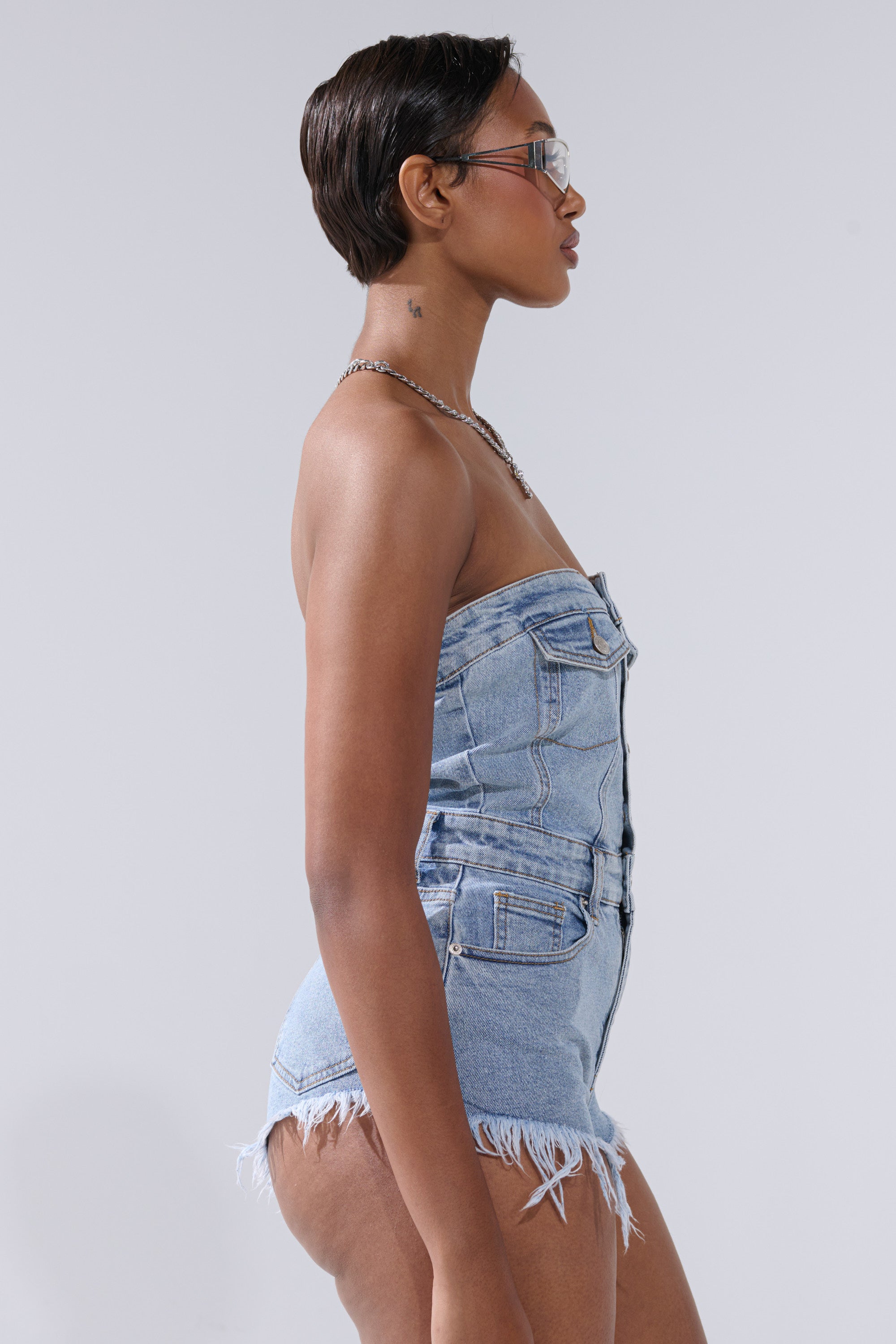 A person with short dark hair and glasses models the COOL IT COWBOY DENIM ROMPER, a strapless western-style romper with frayed shorts, standing in profile against a plain light background.