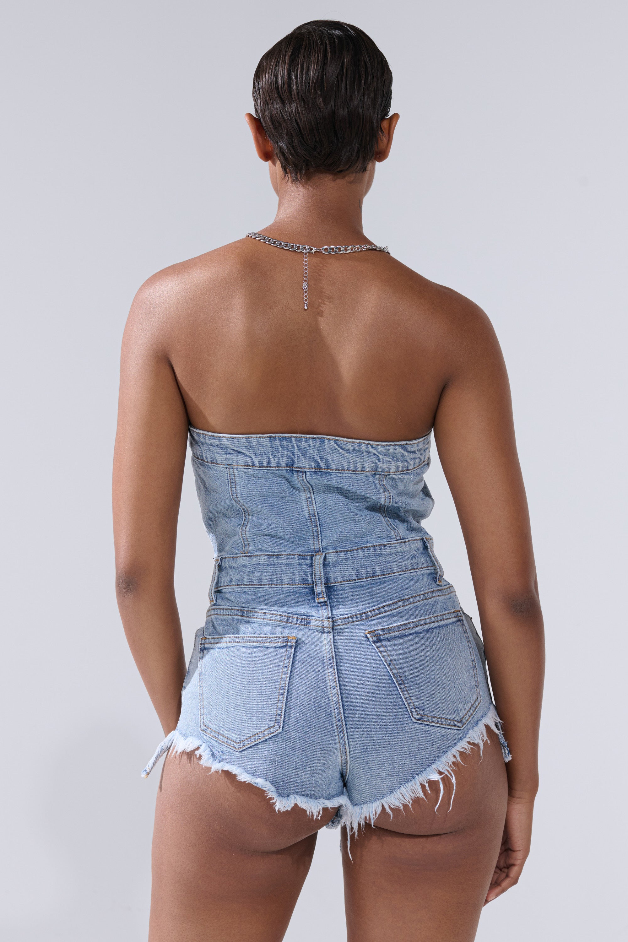 A person with short hair is seen from the back, wearing the COOL IT COWBOY DENIM ROMPER featuring frayed shorts and a silver chain necklace, channeling a western vibe against a plain light gray background.