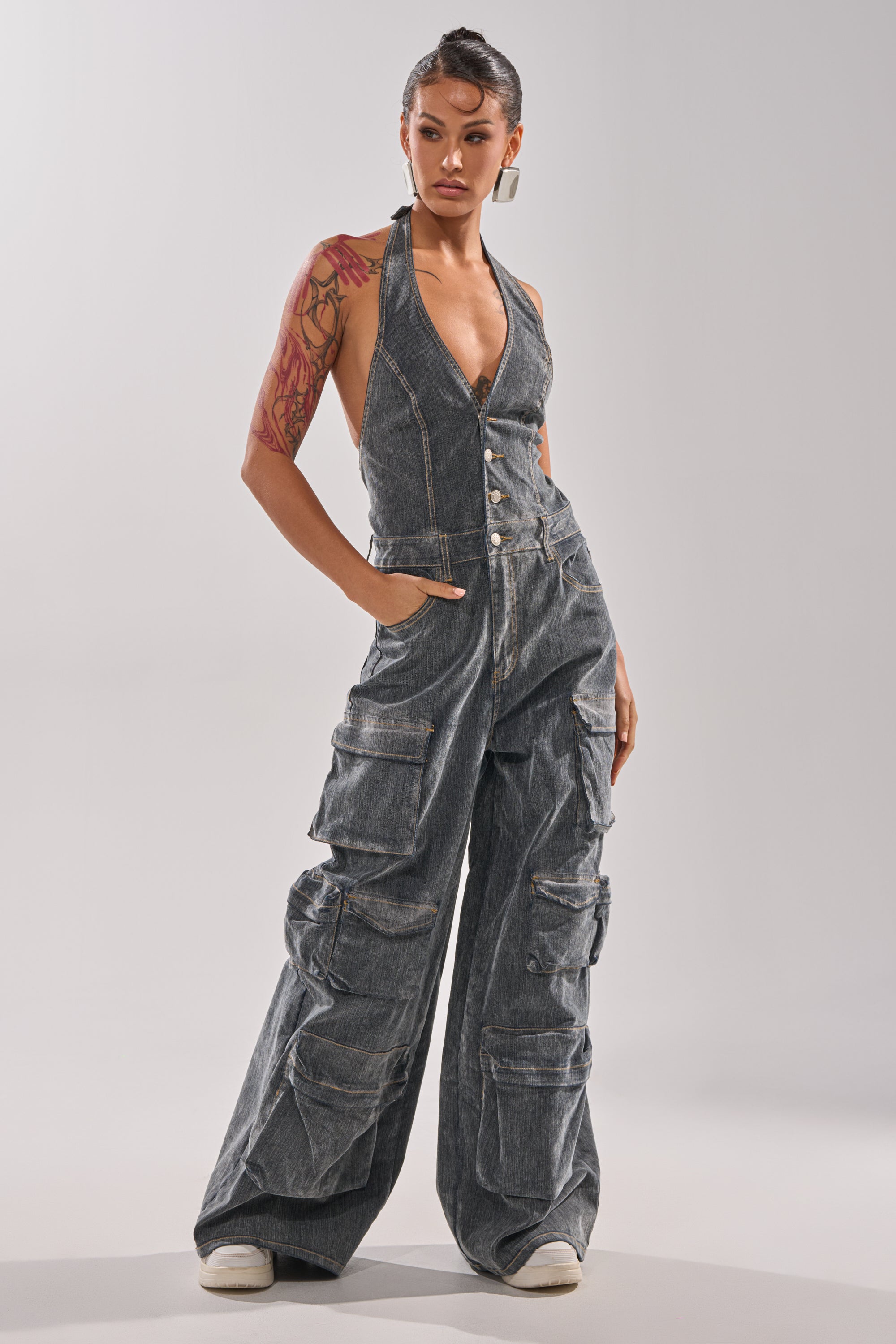 A person stands confidently with hands in pockets, wearing the MELINDA VELVET JUMPSUIT—sleeveless, deep V-neck, wide-leg style with multiple cargo pockets—paired with hoop earrings, white sneakers, and arm tattoos visible. 0