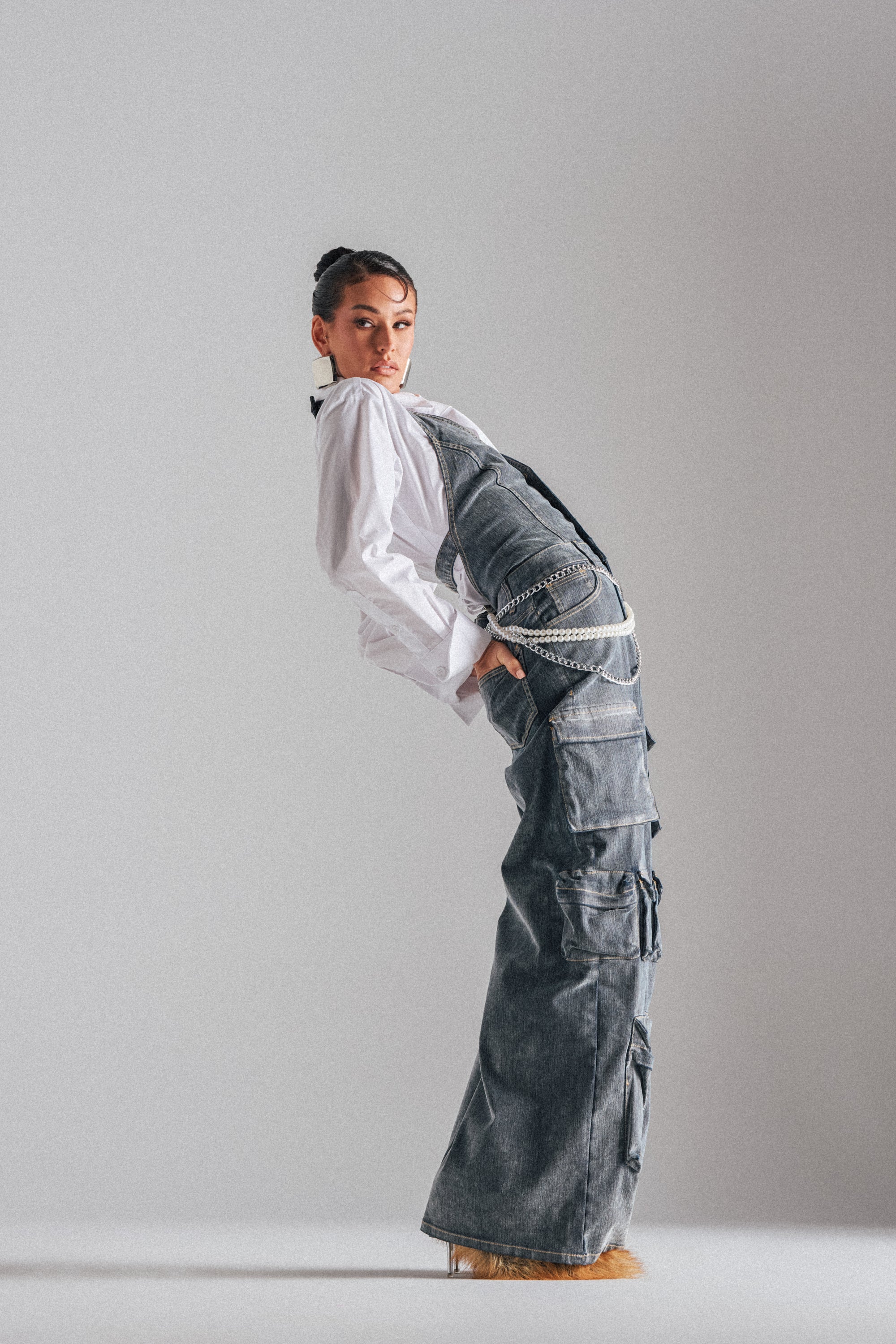 A person in a white shirt and the MELINDA VELVET JUMPSUIT poses dramatically with hands in pockets, leaning back against a plain light gray background. 1