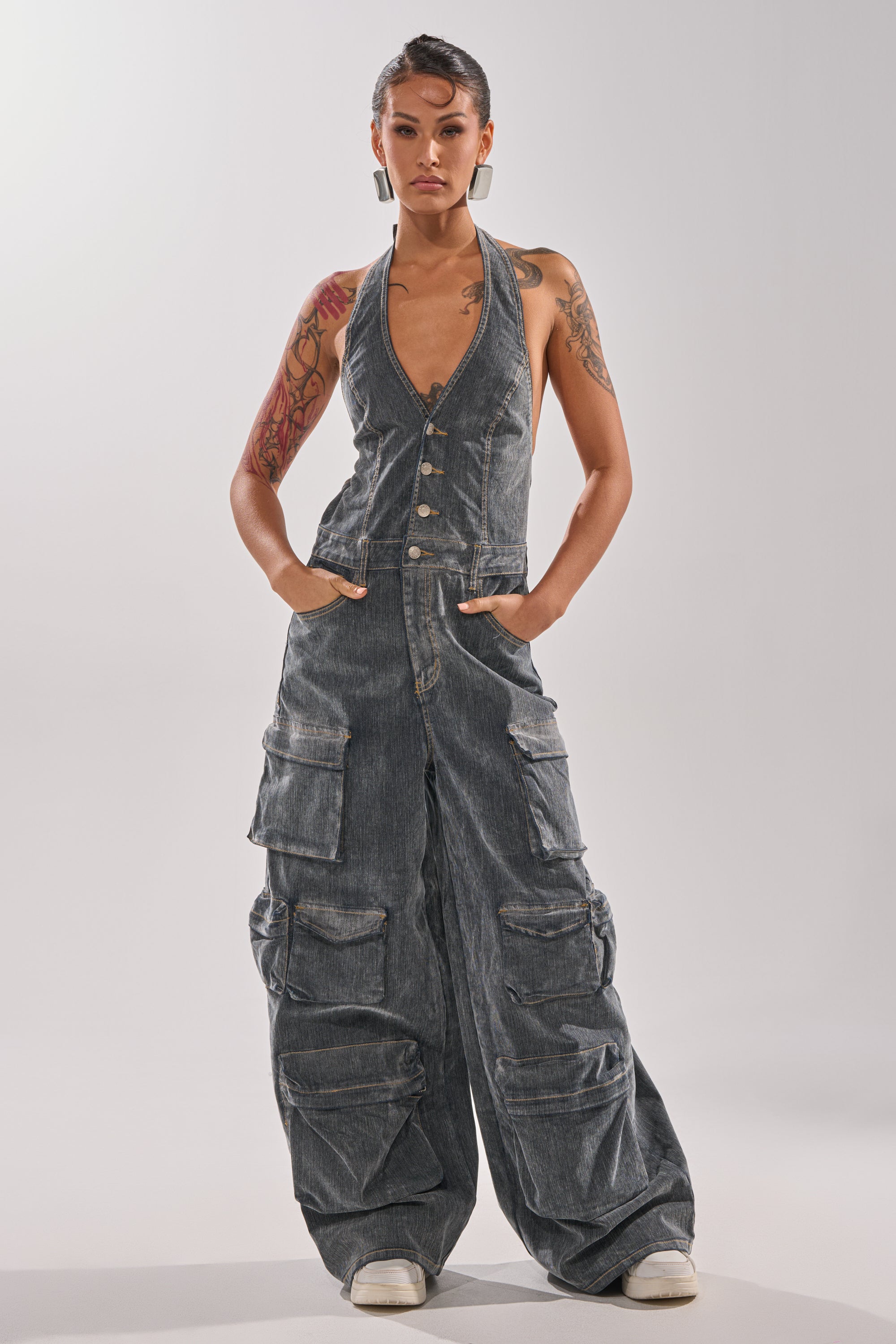 A person stands confidently with hands in pockets, wearing the MELINDA VELVET JUMPSUIT—a sleeveless, deep V-neck wide-leg style—paired with white sneakers and showcasing visible tattoos on their arms. 4