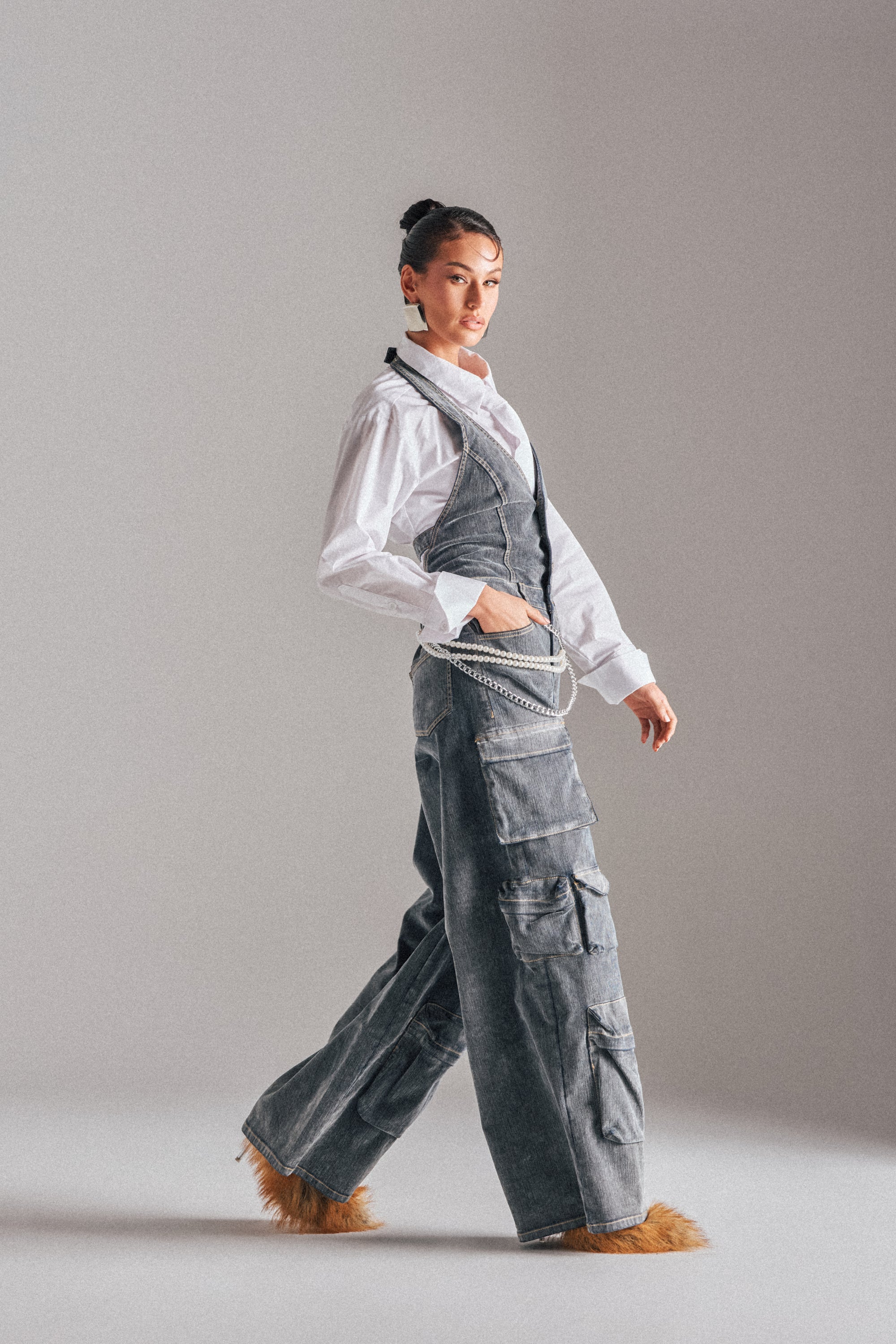 A person with dark hair in a bun wears the MELINDA VELVET JUMPSUIT layered over a white shirt, cargo pants with chain accents, and faux fur shoes, standing against a plain light background. 2