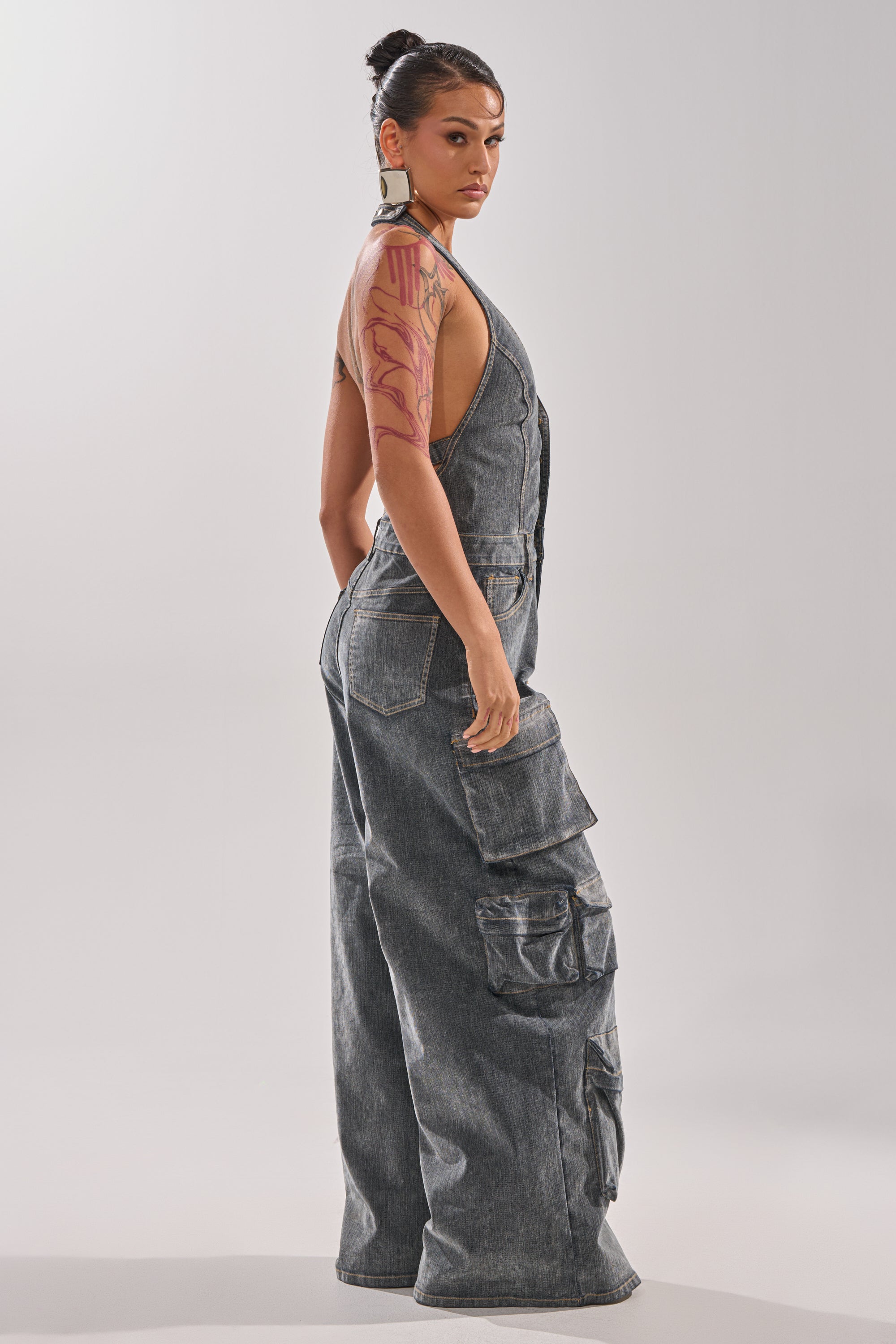 A person with dark hair in a bun, large earrings, and a visible arm tattoo stands in profile against a plain light background, wearing the loose-fitting MELINDA VELVET JUMPSUIT with side pockets. 5
