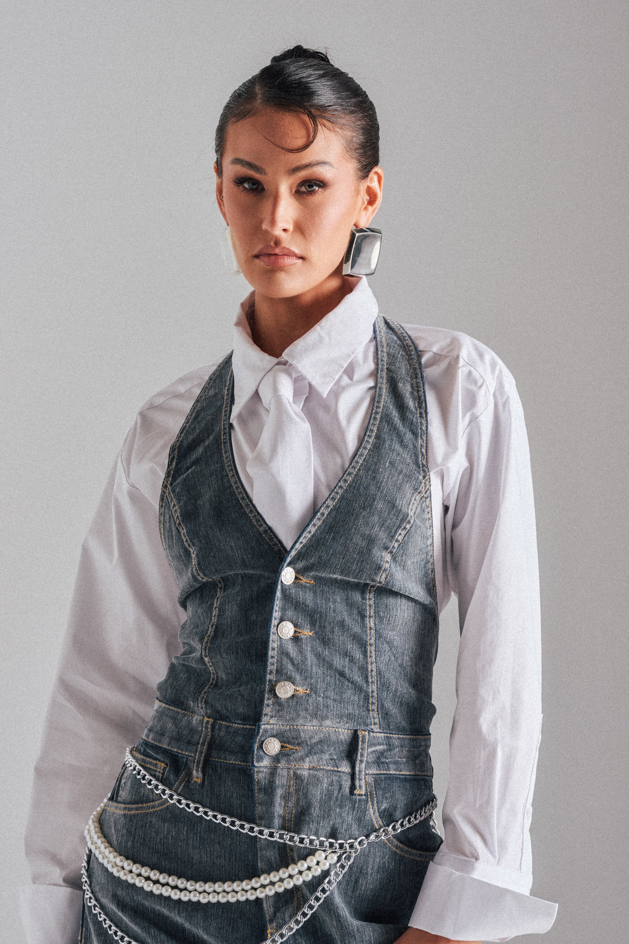 A woman styles the MELINDA VELVET JUMPSUIT with a chunky chain belt and large square earrings, creating a chic look accented by bold accessories and an updo. 3