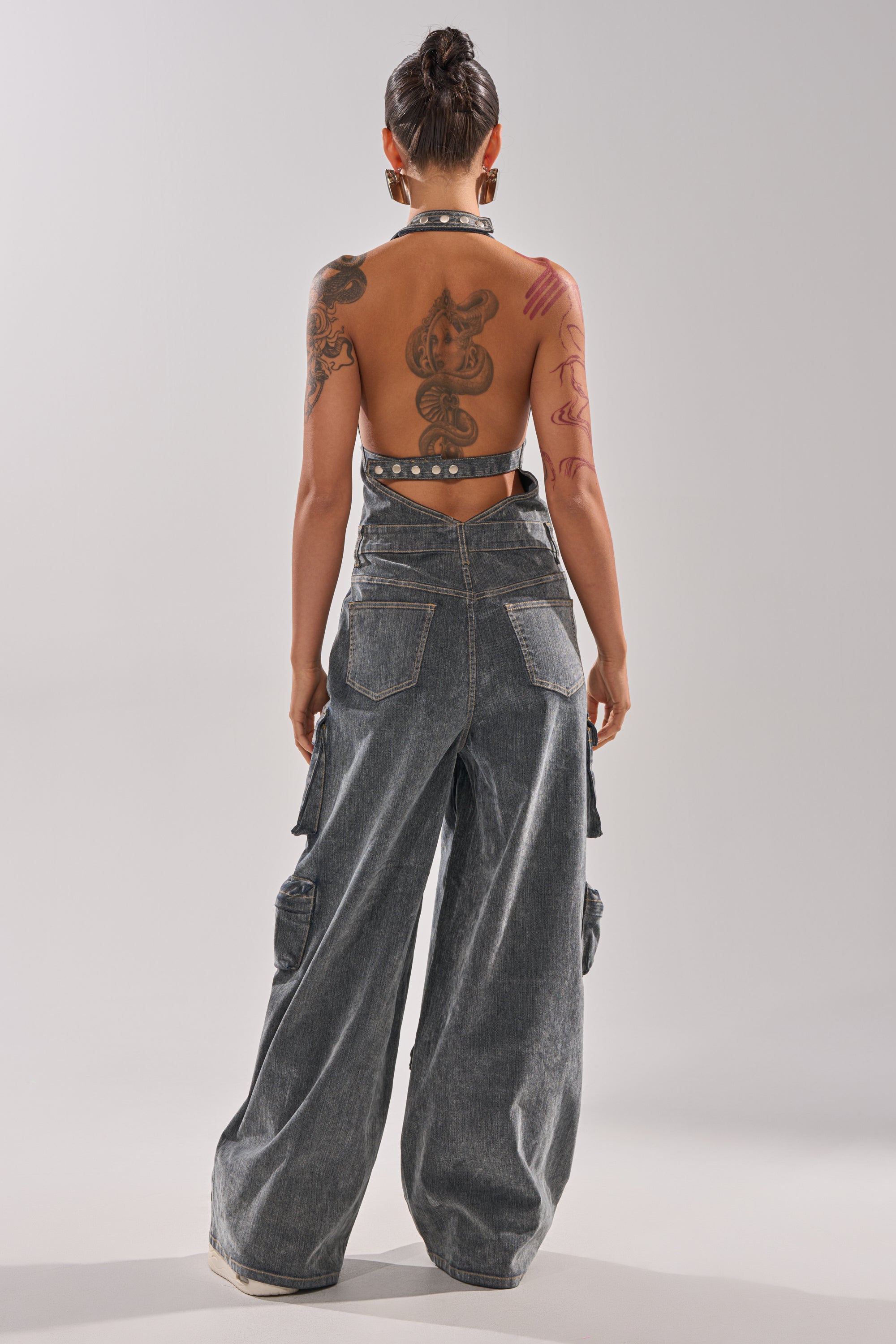 A tattooed individual wearing the MELINDA VELVET JUMPSUIT with a backless halter top and wide-leg fit stands facing away from the camera against a plain, light gray background. 6