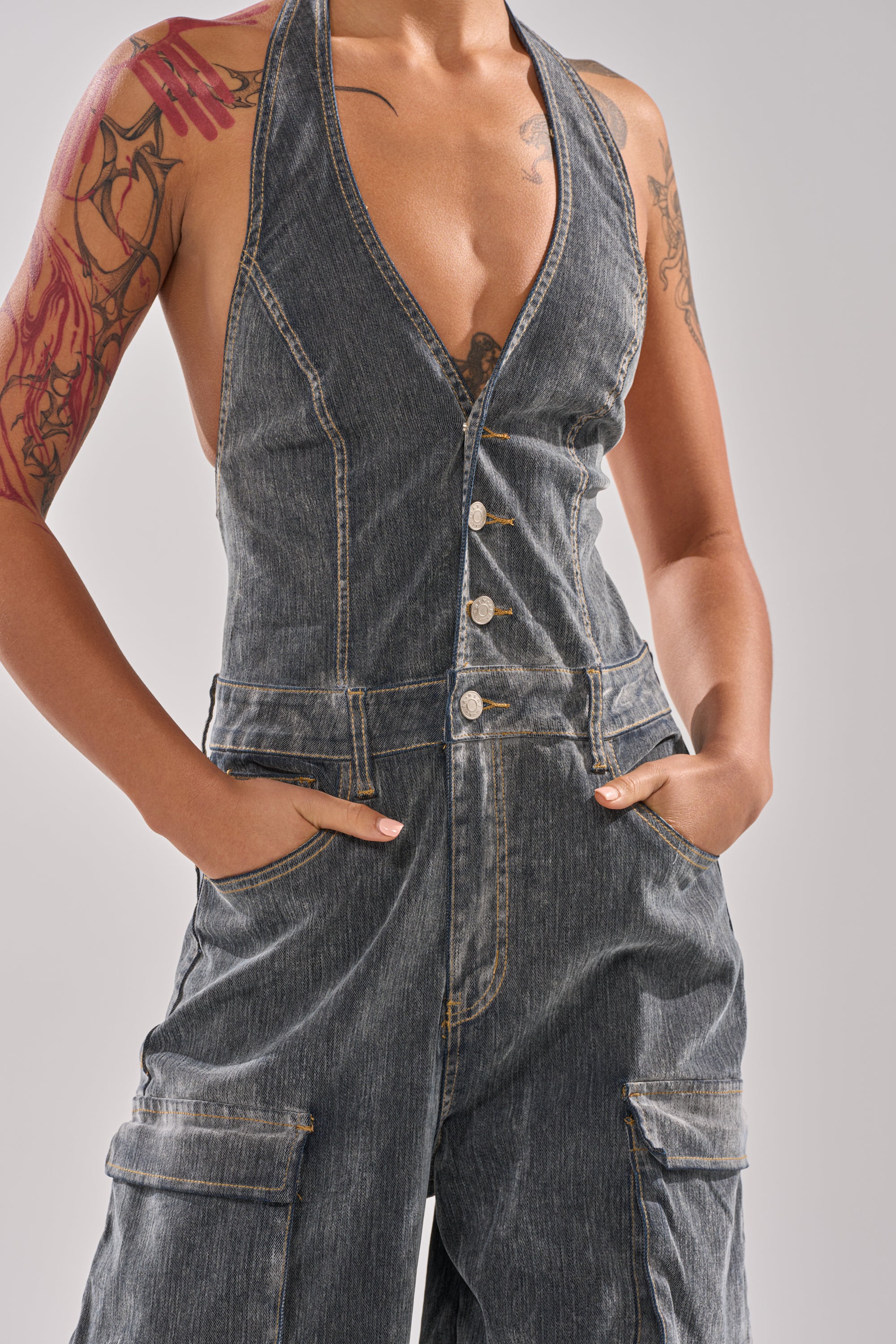 A person models the MELINDA VELVET JUMPSUIT, a sleeveless wide-leg suit with a deep V-neck, metal buttons, and cargo pockets; their tattoos are visible on their arms and shoulders as they pose with hands in pockets. 7
