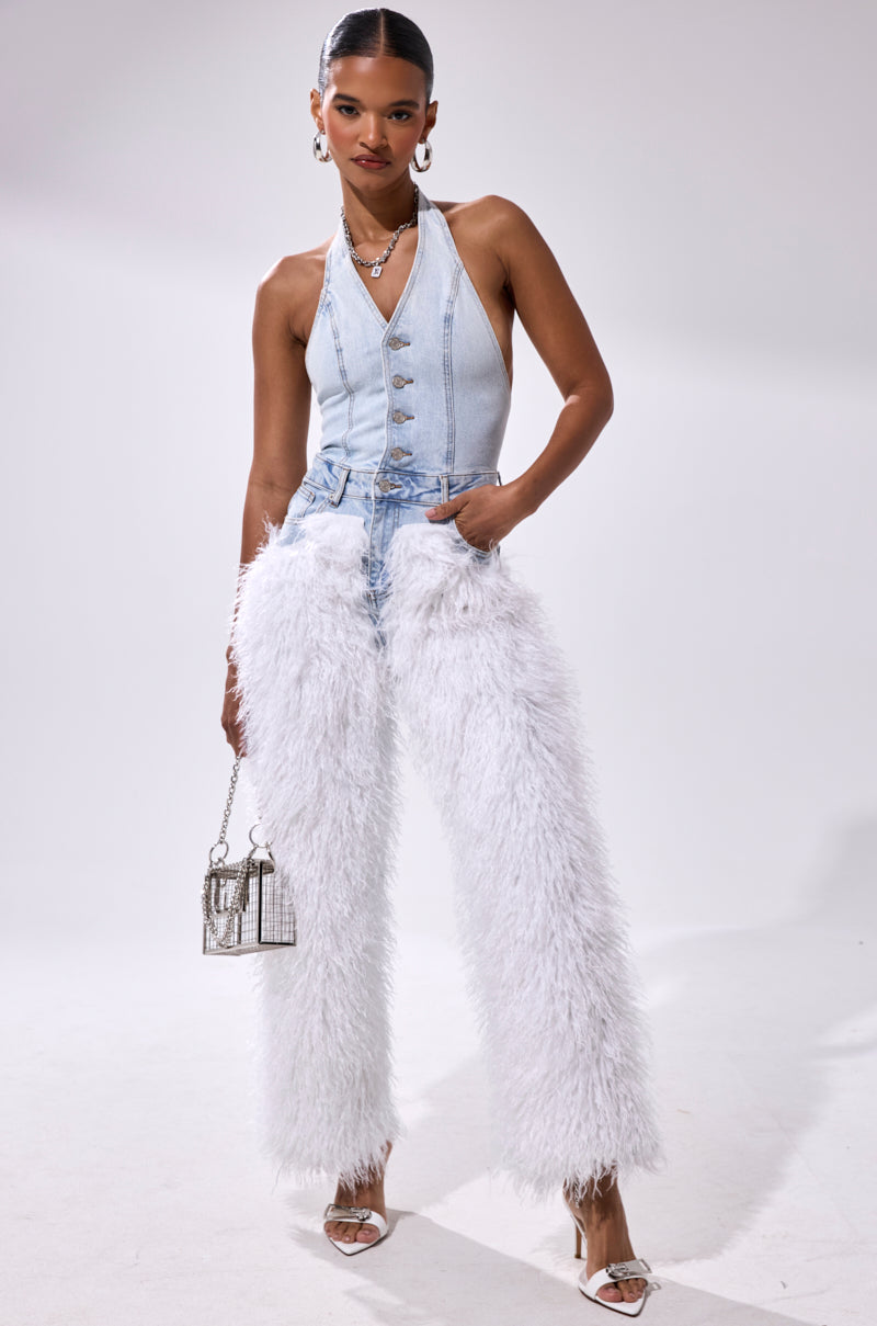 A woman poses in the BADDIES ONLY DENIM FUR JUMPSUIT, a Y2K-inspired sleeveless denim halter style with silver buttons and white furry high-waist details, styled with silver jewelry, a metallic purse, and white strappy heels.