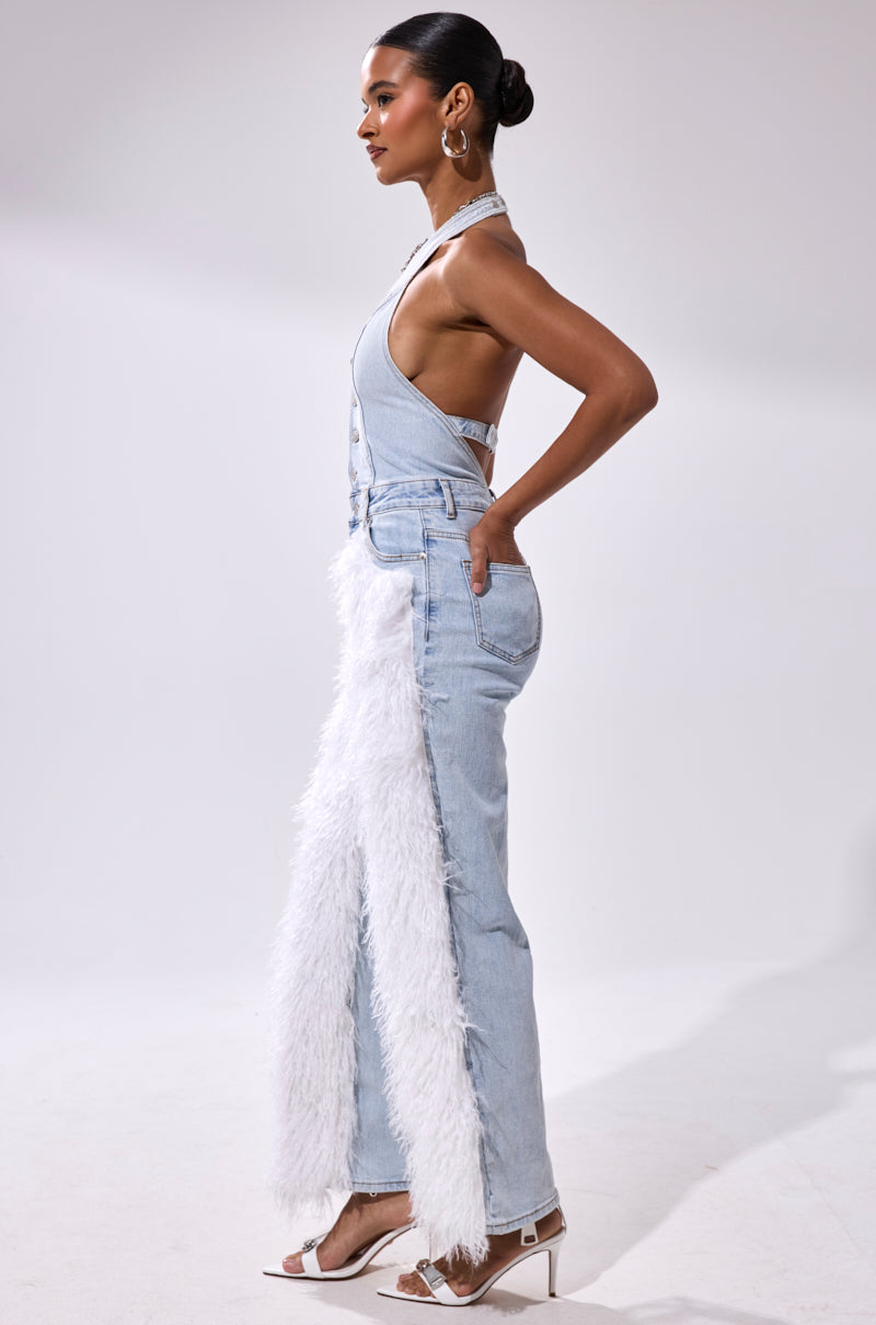 A woman in profile models the BADDIES ONLY DENIM FUR JUMPSUIT, featuring an open back, wide-leg pants, faux fur trim on one leg, paired with hoop earrings and white high-heeled sandals against a plain backdrop.
