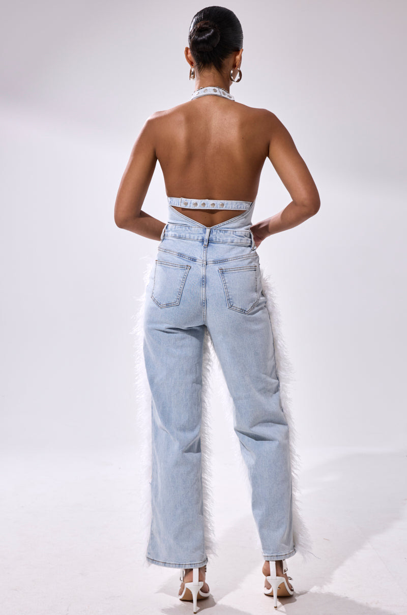 A woman, hair in a neat bun, models the BADDIES ONLY DENIM FUR JUMPSUIT—a Y2K-inspired piece featuring light blue high-waisted wide-leg jeans with white feather trim and a backless halter top—paired with white heels.