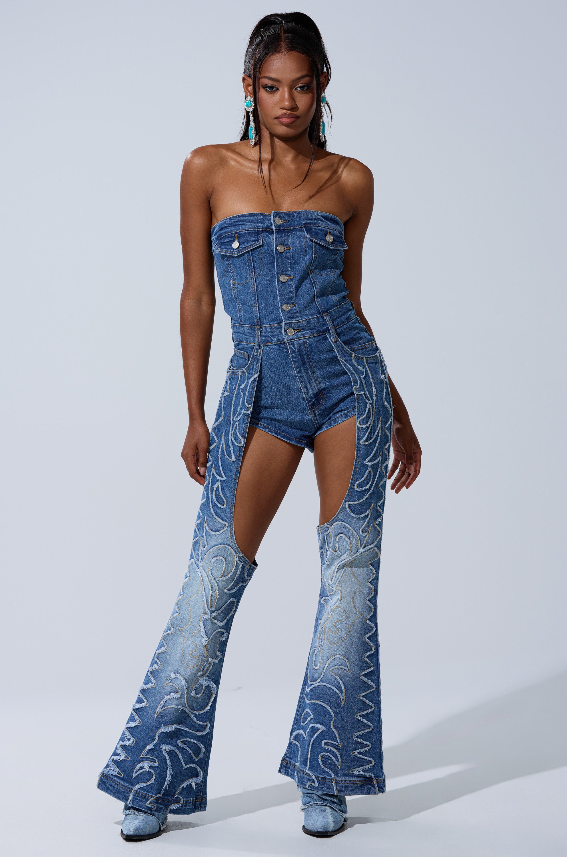 A woman models the THROW DOWN DENIM JUMPSUIT WITH CHAPS, a western-inspired strapless button-up outfit with matching flared chaps featuring light blue embroidery, styled with denim boots and turquoise earrings.