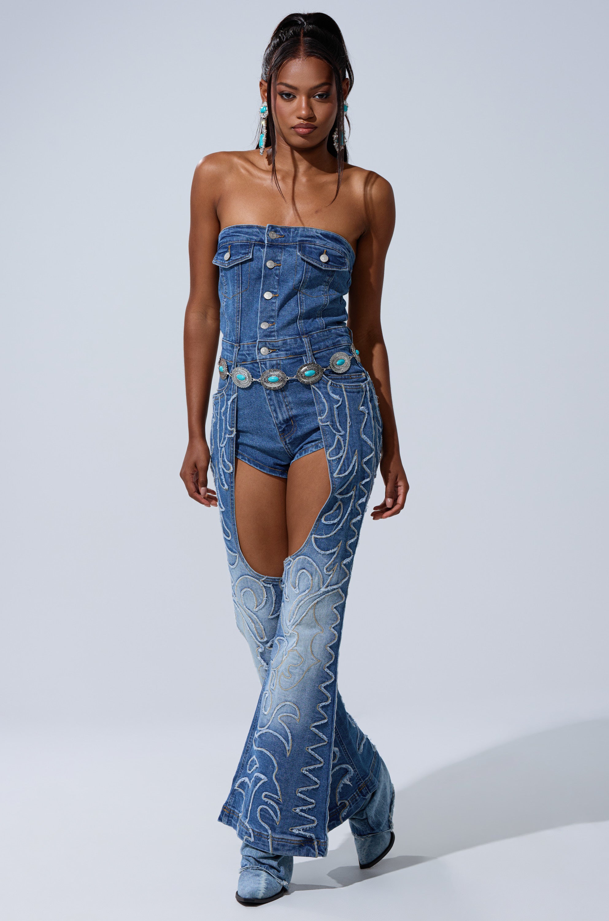 A woman wearing the THROW DOWN DENIM JUMPSUIT WITH CHAPS, featuring western-inspired embroidery, turquoise accents, and blue boots, walks confidently against a plain background.