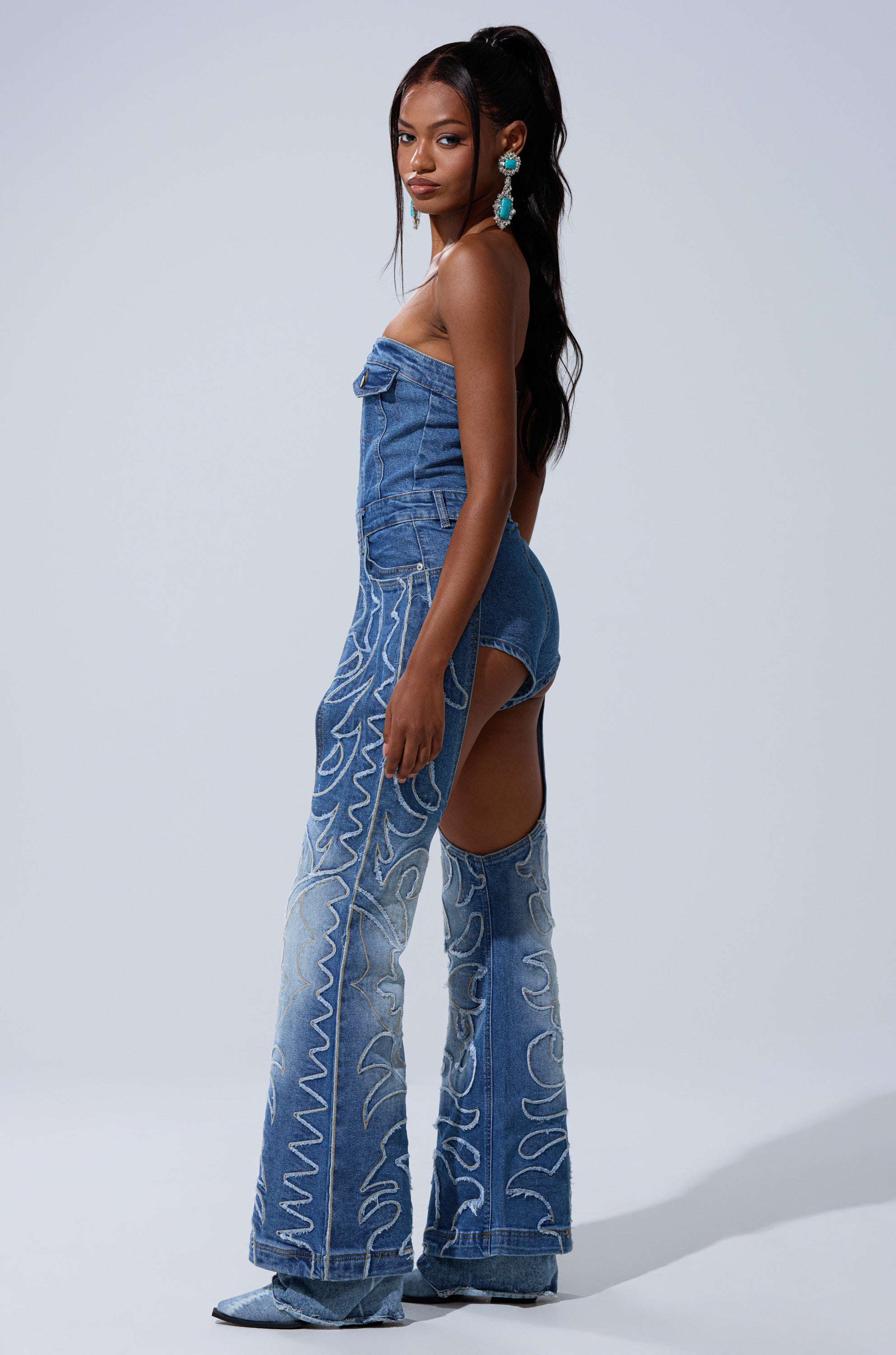 A woman poses sideways against a plain light background in the THROW DOWN DENIM JUMPSUIT WITH CHAPS, featuring hip cutouts and swirling white stitching. She accessorizes with statement earrings and wears her long hair styled.