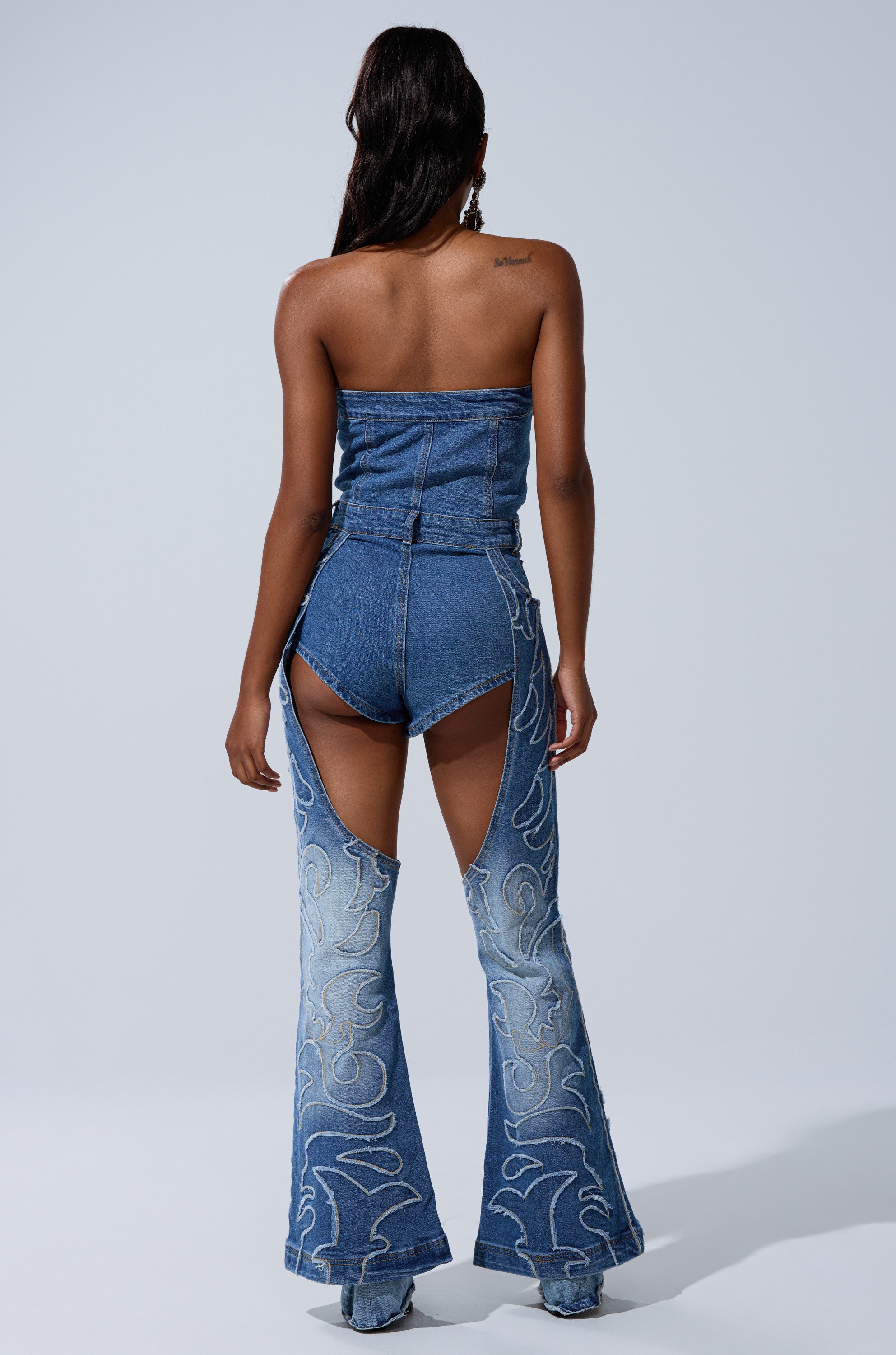 A woman models the THROW DOWN DENIM JUMPSUIT WITH CHAPS, a strapless, western-inspired outfit featuring chap-style pants and light blue embroidered swirls, exposing her arms, back, and upper thighs.