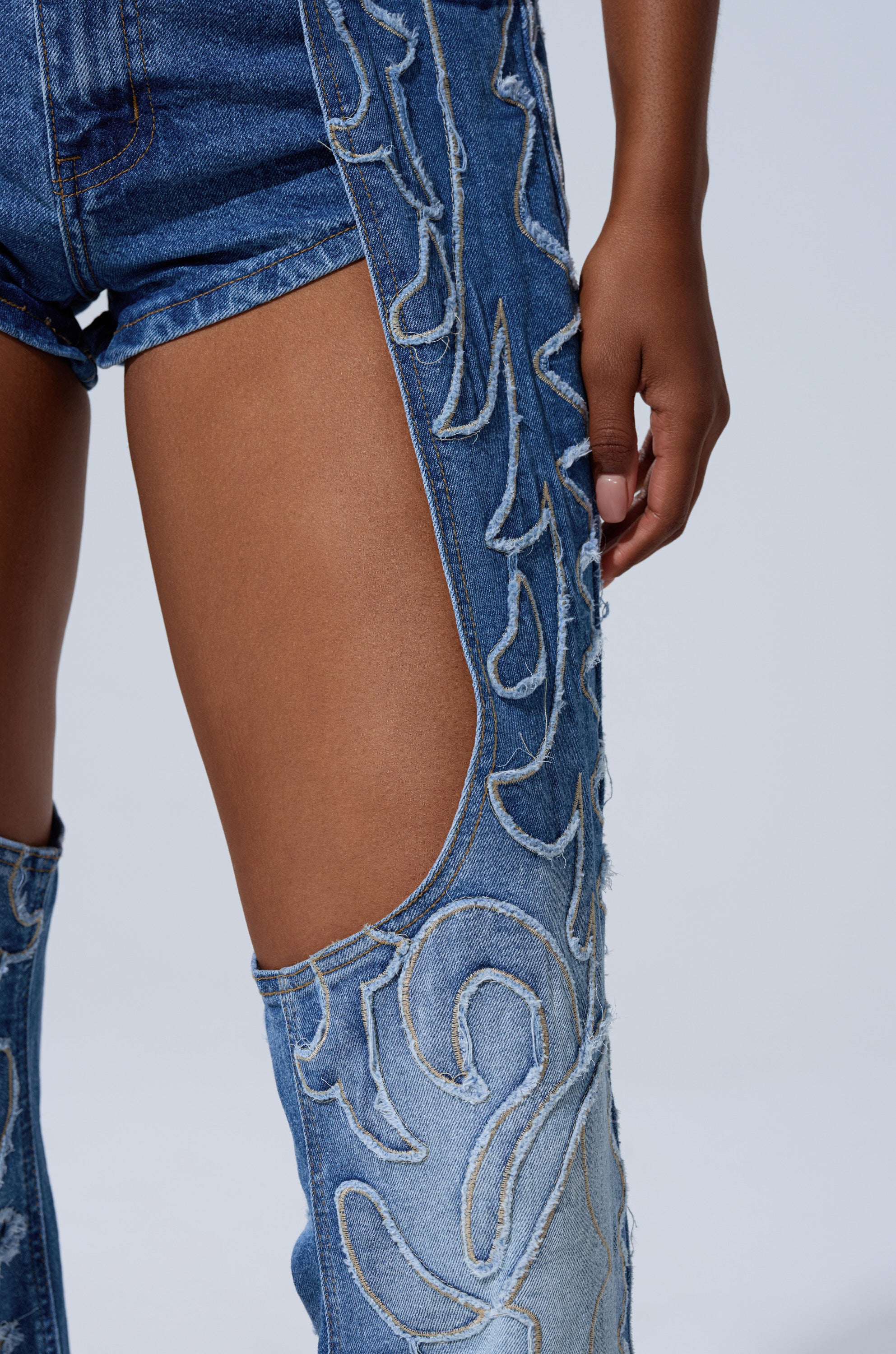 A person wears the THROW DOWN DENIM JUMPSUIT WITH CHAPS, featuring chap-style denim shorts and matching thigh-high boots with swirling stitched patterns and open thigh sections. Their hand rests at their side.