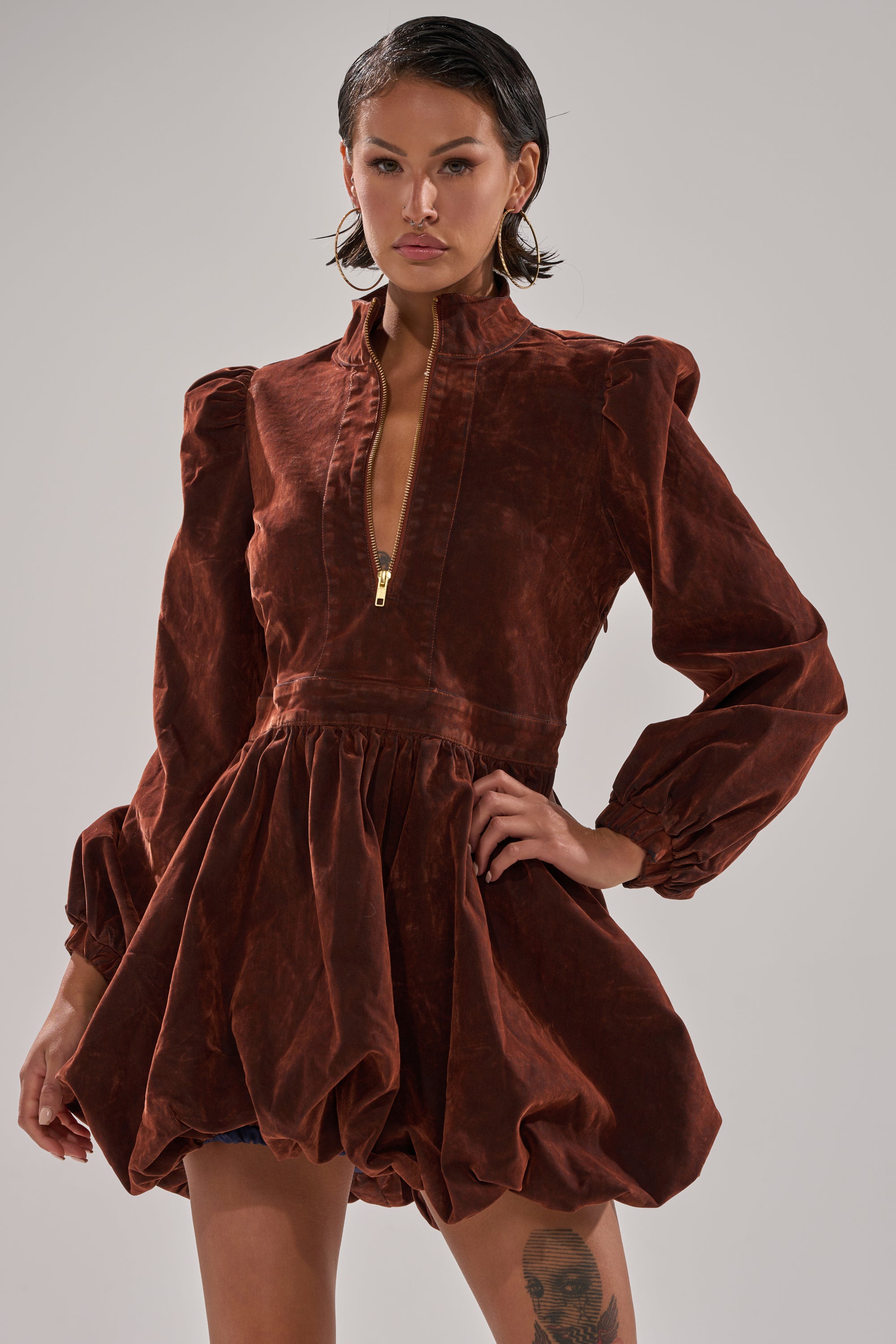 A woman stands confidently with one hand on her hip, modeling the PASS THE BUBBLY MINI DRESS IN VELVET DENIM—a rust-colored bubble hem mini dress with long puffed sleeves, a high collar, and flared skirt, set against a plain background.