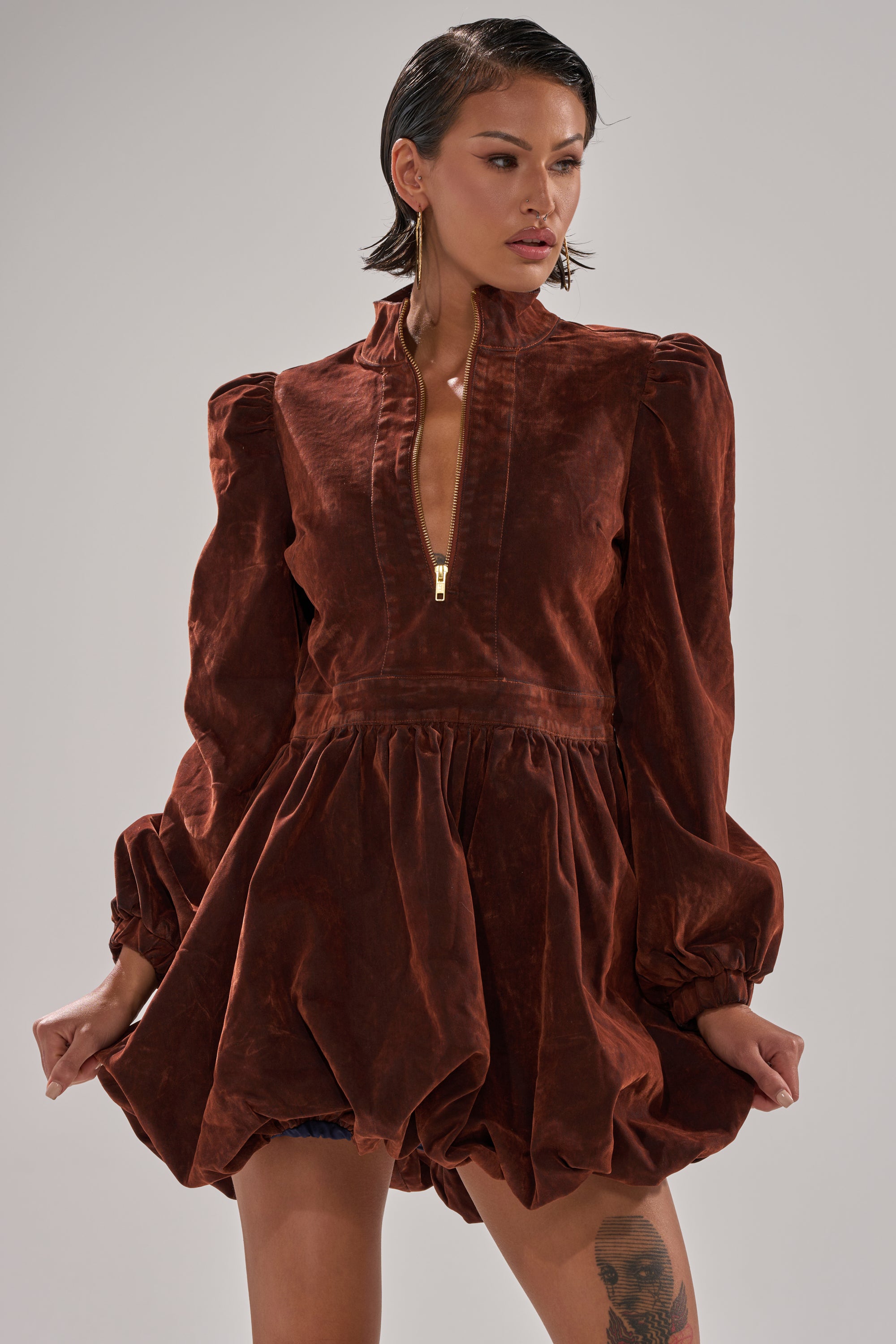 A person wears the PASS THE BUBBLY MINI DRESS IN VELVET DENIM, featuring puff sleeves, a bubble hem, front zipper, and voluminous skirt. They accessorize with hoop earrings, have slicked-back hair, and display a visible leg tattoo.