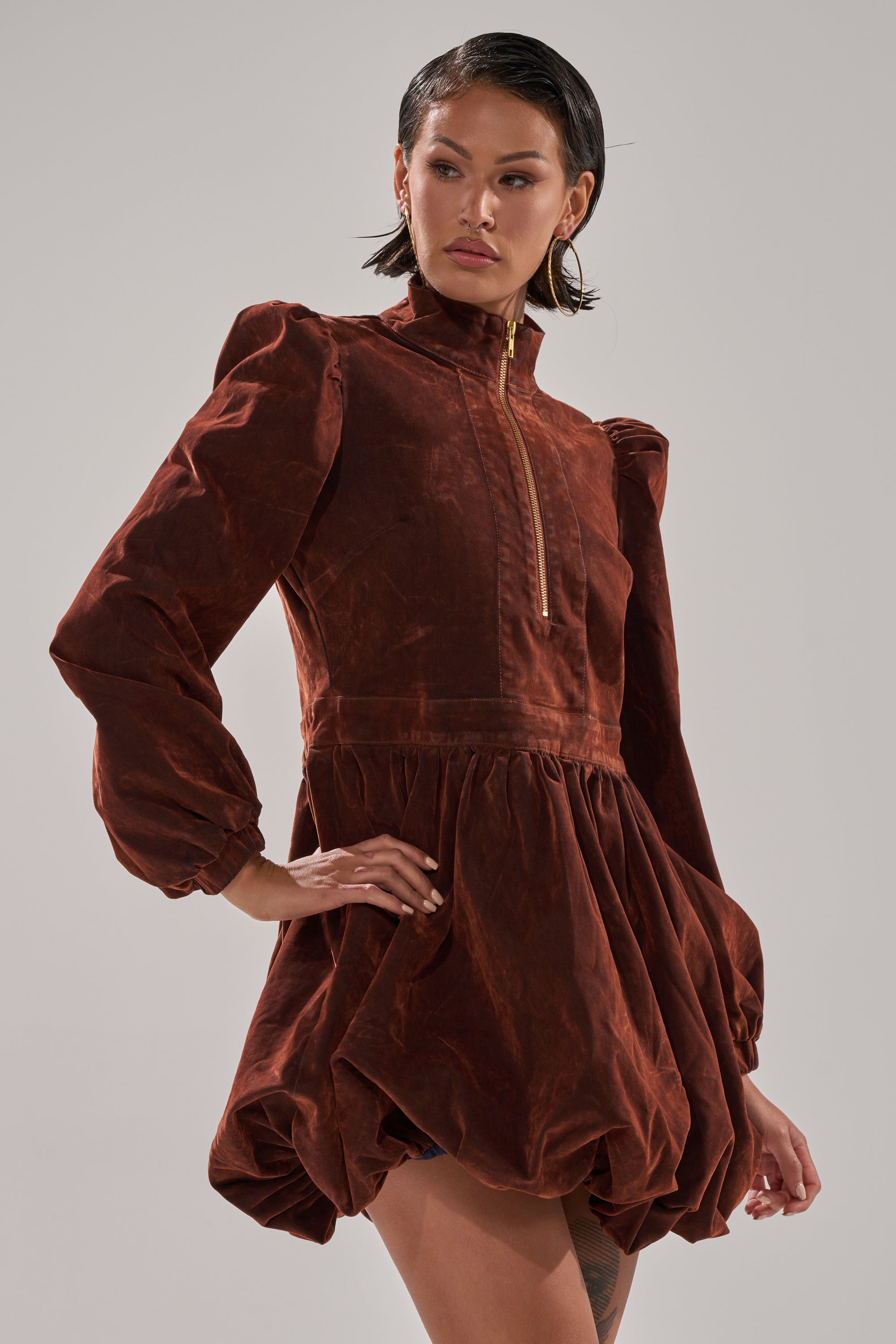 A person poses confidently with one hand on their hip, wearing the PASS THE BUBBLY MINI DRESS IN VELVET DENIM—a rust-colored mock neck dress with long puffed sleeves and a gathered bubble hem—against a plain gray background.