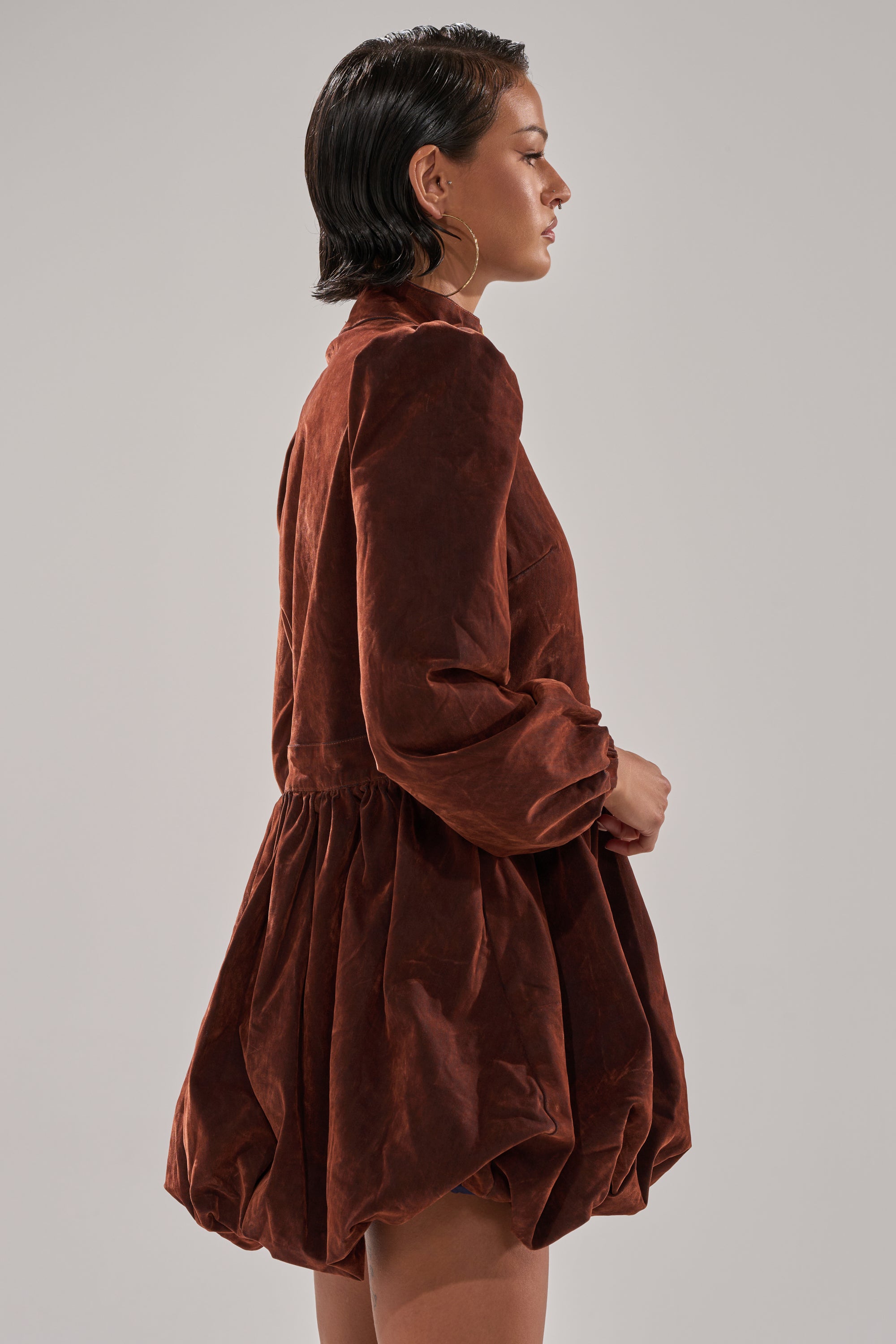 A woman with short dark hair wears the PASS THE BUBBLY MINI DRESS IN VELVET DENIM, a brown, long-sleeved mock neck dress with a voluminous bubble hem skirt. She stands sideways against a plain gray background.