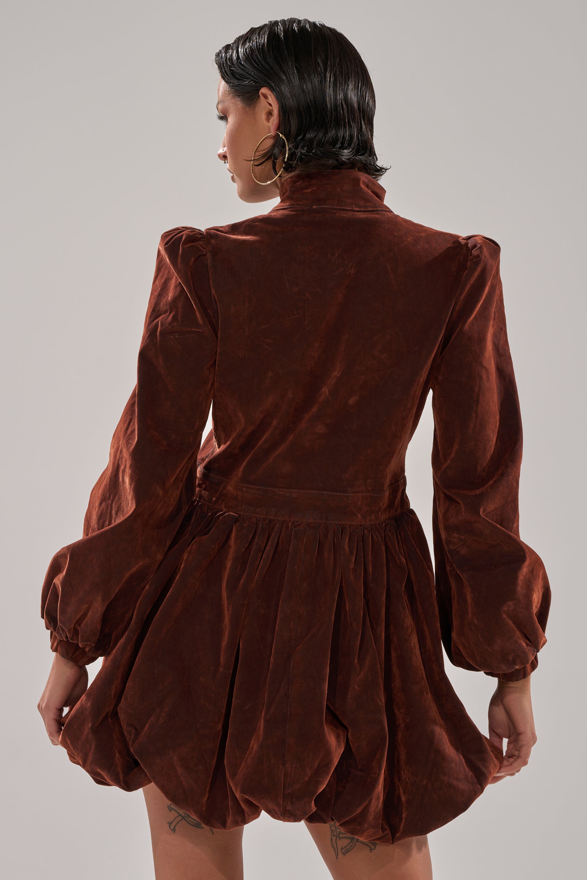 A person with short, wet-look hair and hoop earrings is shown from the back wearing the PASS THE BUBBLY MINI DRESS IN VELVET DENIM, featuring long sleeves and a gathered skirt, against a plain gray background.