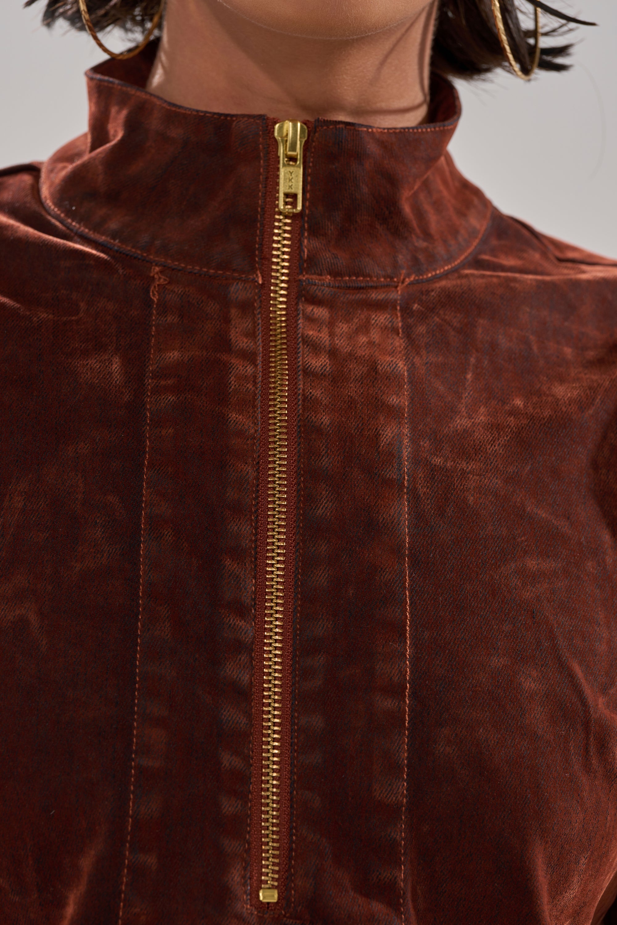 Close-up of a person wearing the PASS THE BUBBLY MINI DRESS IN VELVET DENIM—a brown, high-collared dress with a visible gold center zipper and textured fabric. Only the neck, collar, and part of the chest are shown.