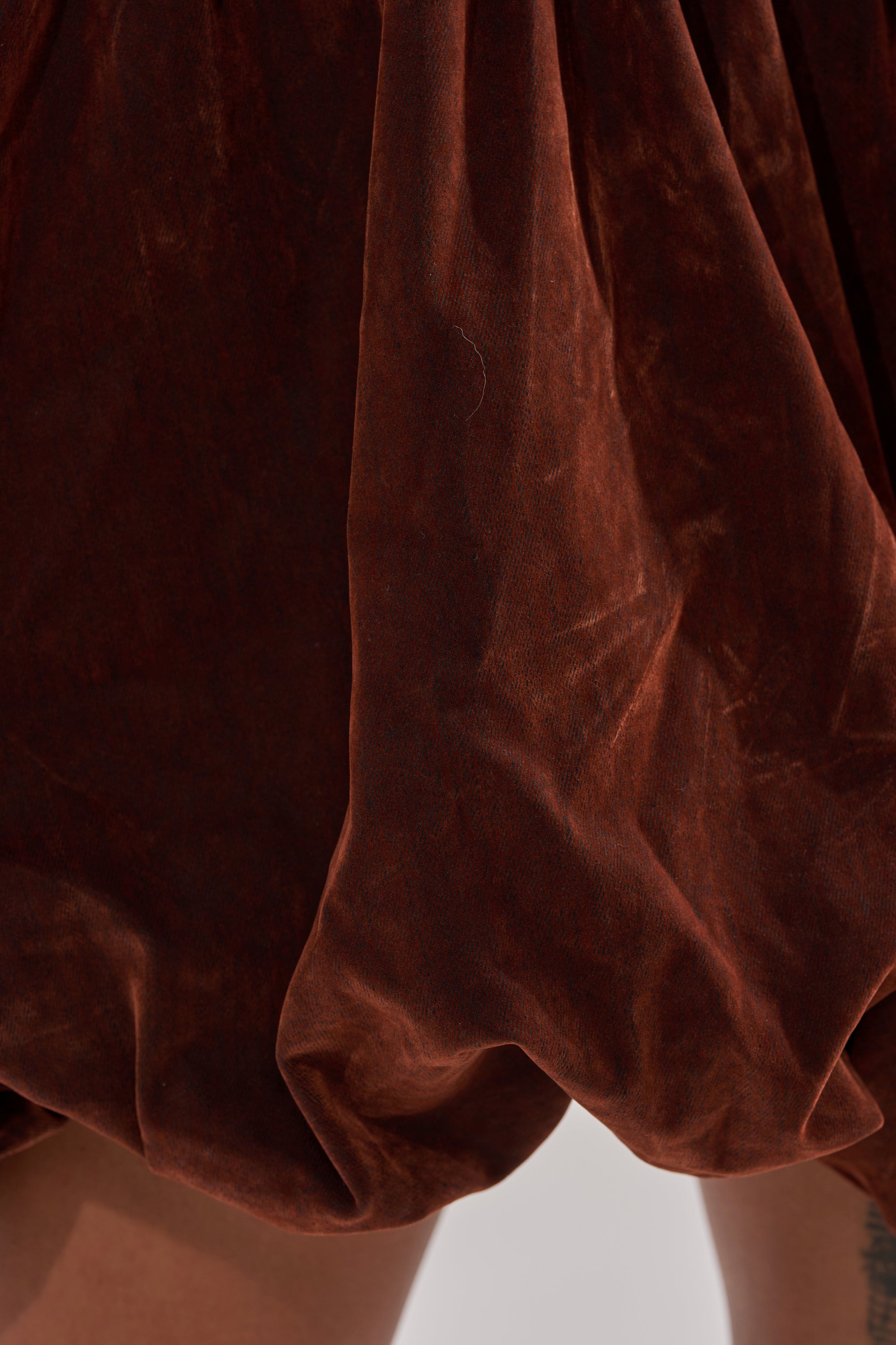 Close-up of the PASS THE BUBBLY MINI DRESS IN VELVET DENIM: rich brown, velvety fabric forms soft folds and catches the light, with a glimpse of a tattooed leg at the bottom right.