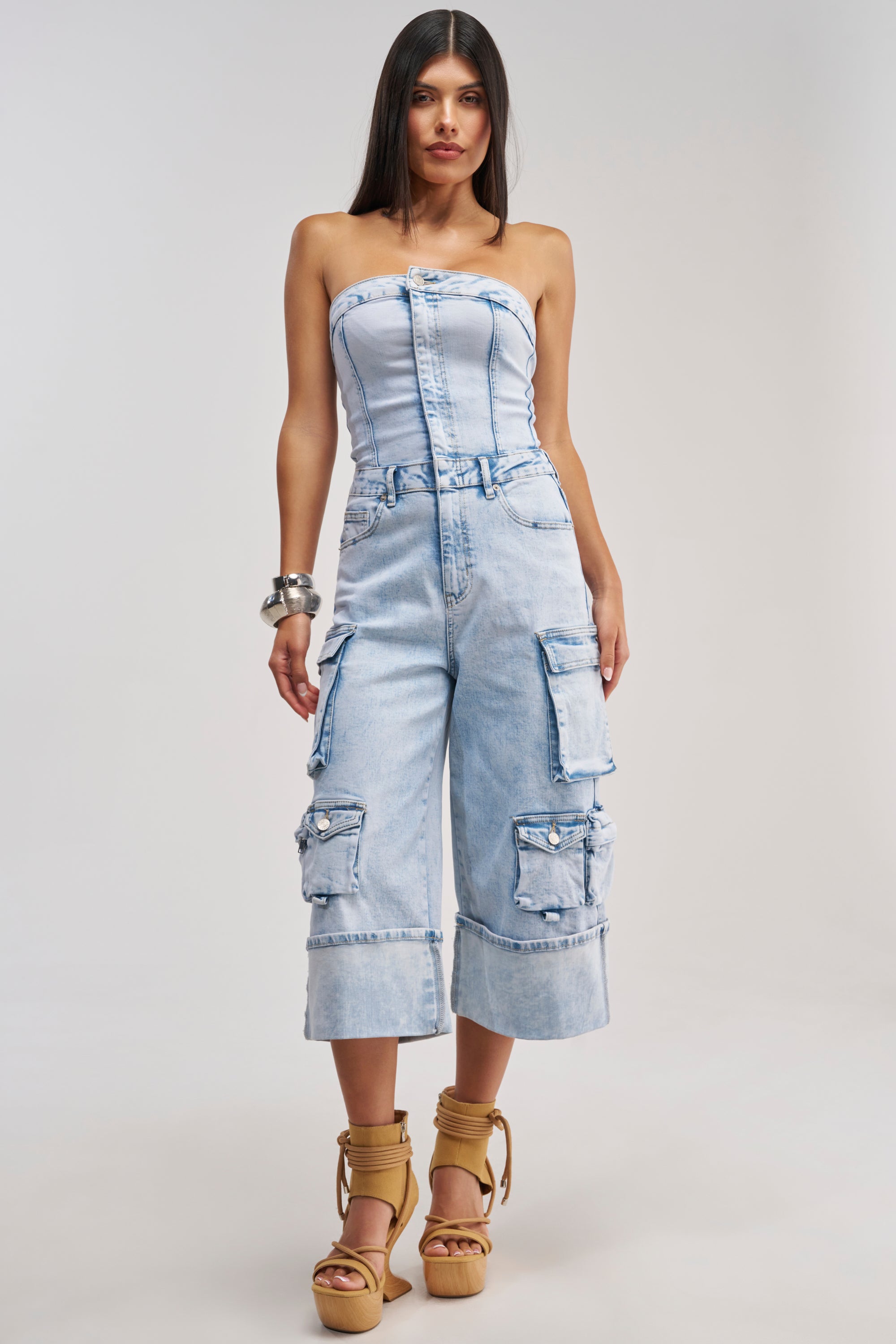A woman stands against a plain light gray background, modeling the ALICIA CROPPED CARGO JUMPSUIT IN LIGHT BLUE DENIM—a strapless design featuring cargo pockets and wide cropped legs that capture a modern streetwear vibe. She accessorizes with long dark hair, tan platform sandals, and silver bangles.