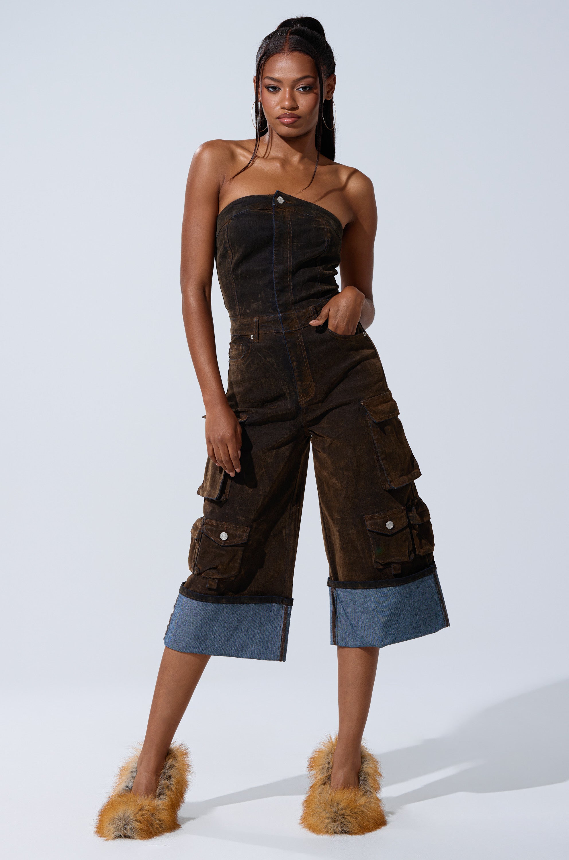 A woman exudes confidence in the ALICIA CROPPED CARGO JUMPSUIT, featuring wide, cuffed legs and denim accents. She pairs it with furry slippers and poses against a plain light background.