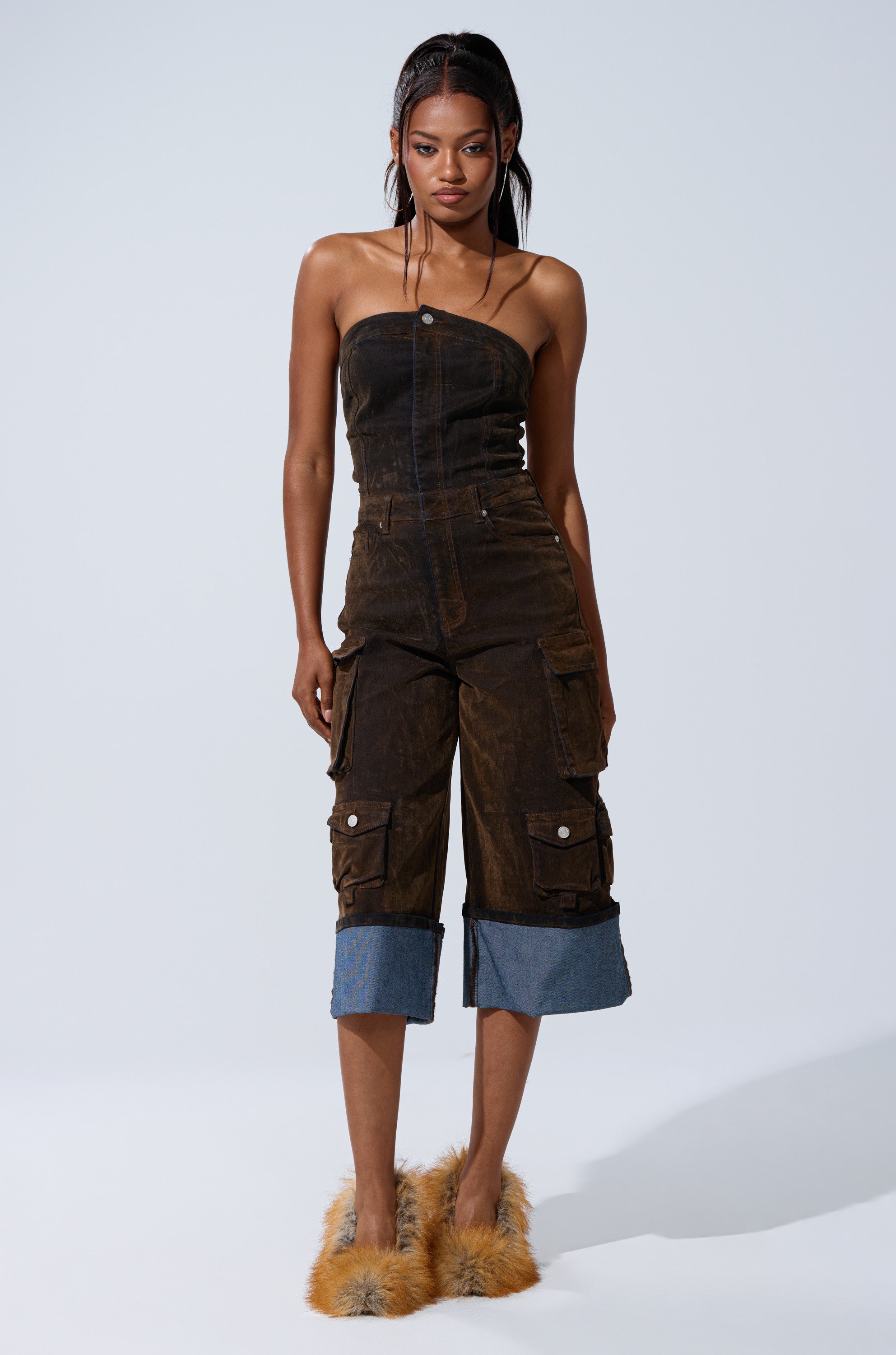A woman with braided hair and a neutral expression stands against a plain background wearing the ALICIA CROPPED CARGO JUMPSUIT, featuring wide legs, cargo pockets, denim cuffs at the calves, and fuzzy tan slippers.