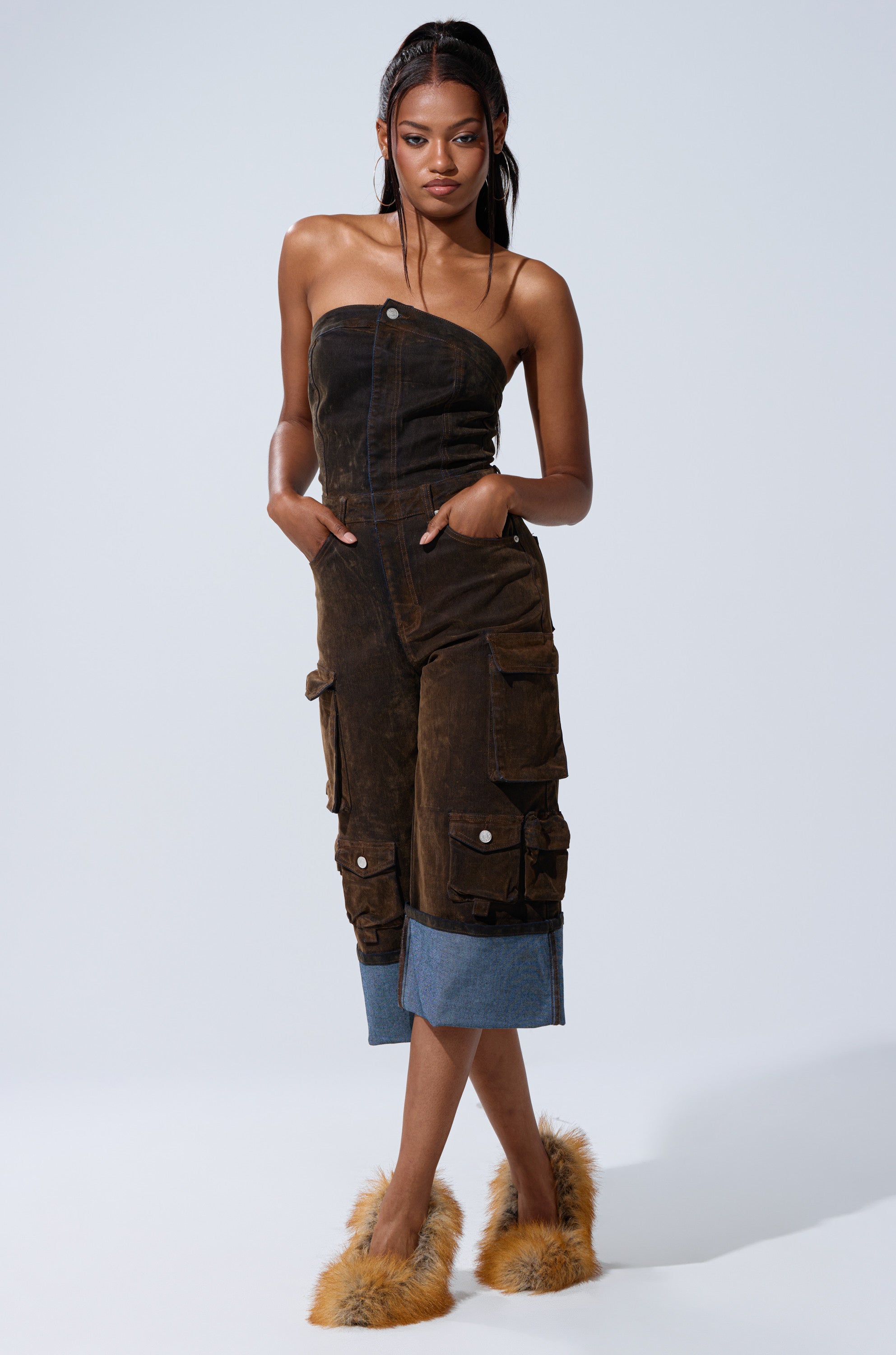 A woman poses against a plain background wearing the ALICIA CROPPED CARGO JUMPSUIT with denim cuffs, her hands in her pockets, and fuzzy brown slippers on her feet.