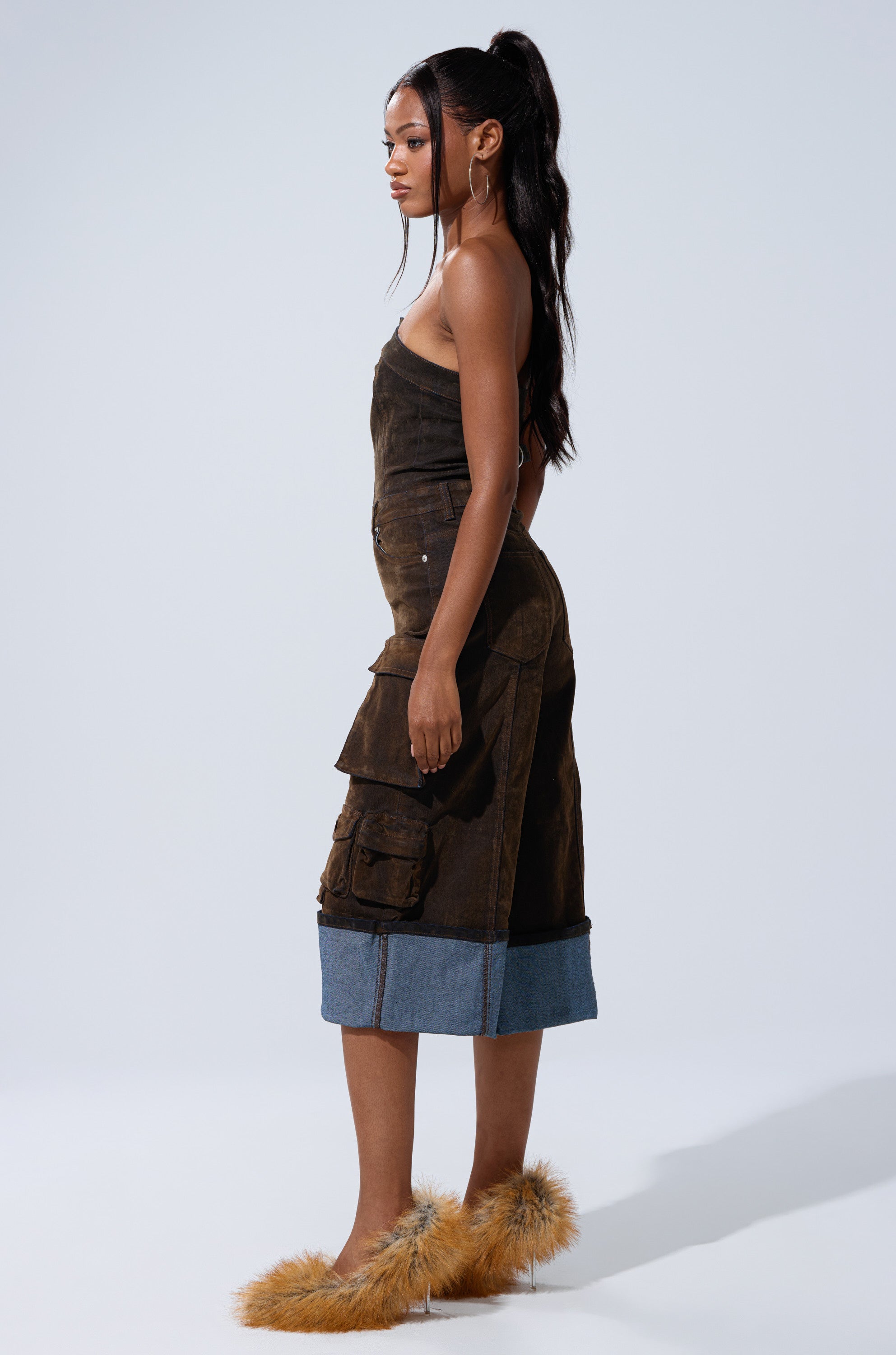 A woman with long dark hair in a ponytail models the ALICIA CROPPED CARGO JUMPSUIT, a brown strapless suede outfit with cargo pockets and denim trim. She stands sideways in tan fluffy heels against a plain light background.
