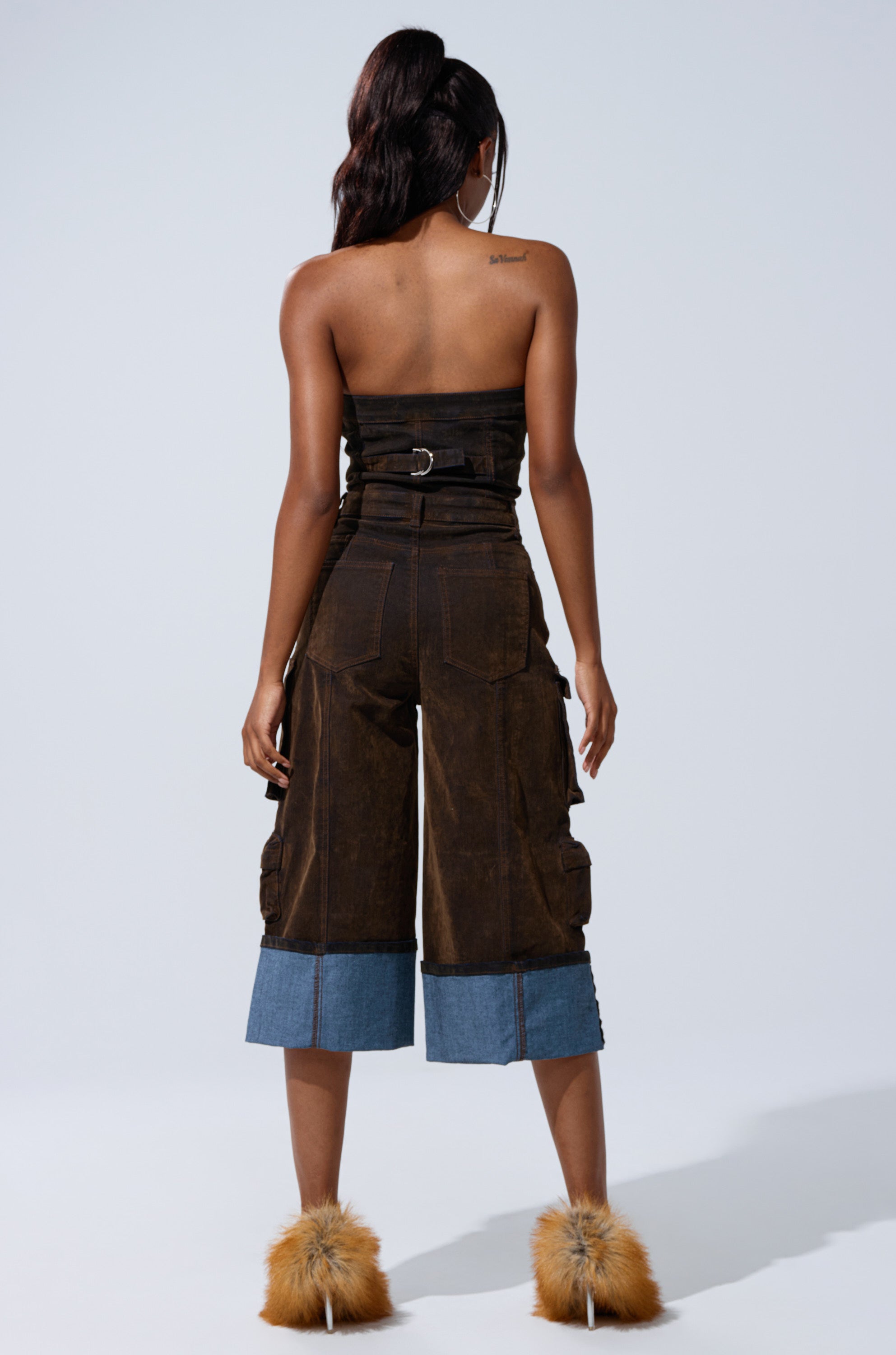 A woman faces away against a plain light background, wearing the ALICIA CROPPED CARGO JUMPSUIT with denim cuffs and fluffy tan open-heeled shoes.