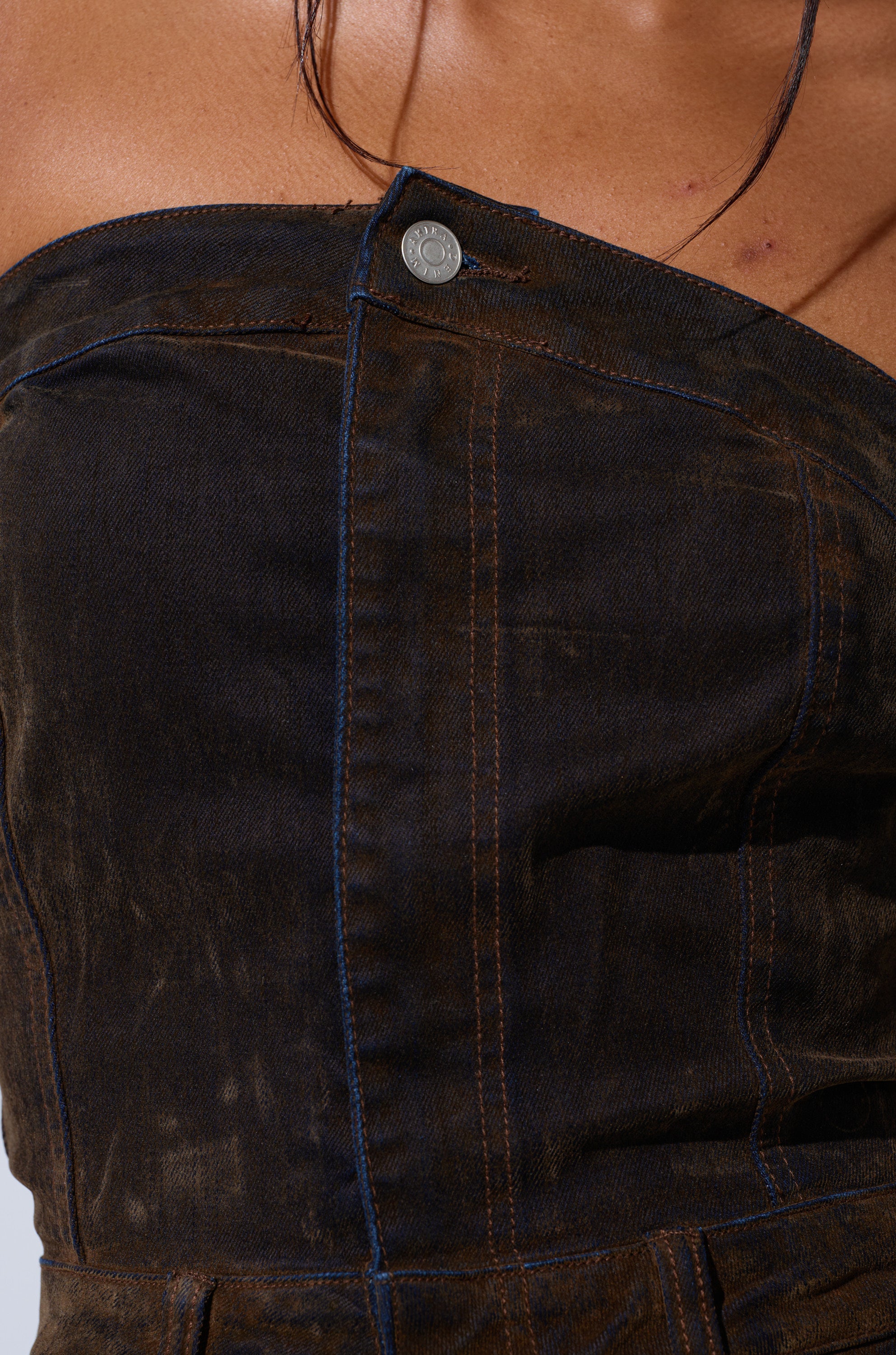 Close-up of a person wearing the ALICIA CROPPED CARGO JUMPSUIT, featuring dark denim, an asymmetrical strapless neckline, visible brown stitching, and a metal button near the collar. Strands of hair fall across their bare shoulder and neck.