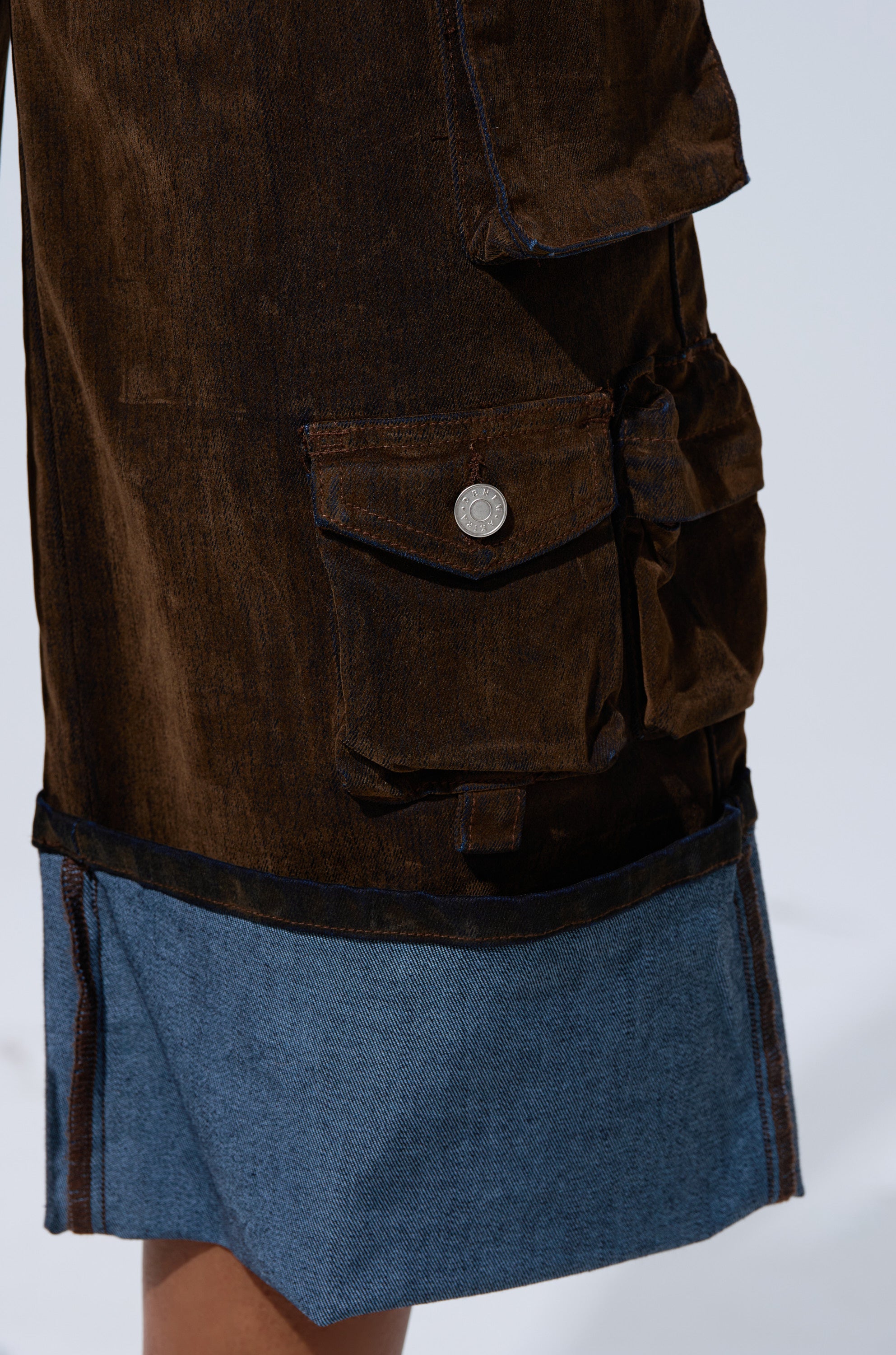 Close-up of the ALICIA CROPPED CARGO JUMPSUIT showcases brown denim with a large side pocket, button closure, and wide blue hem—featuring visible fabric texture and stitching for a utilitarian-inspired look.