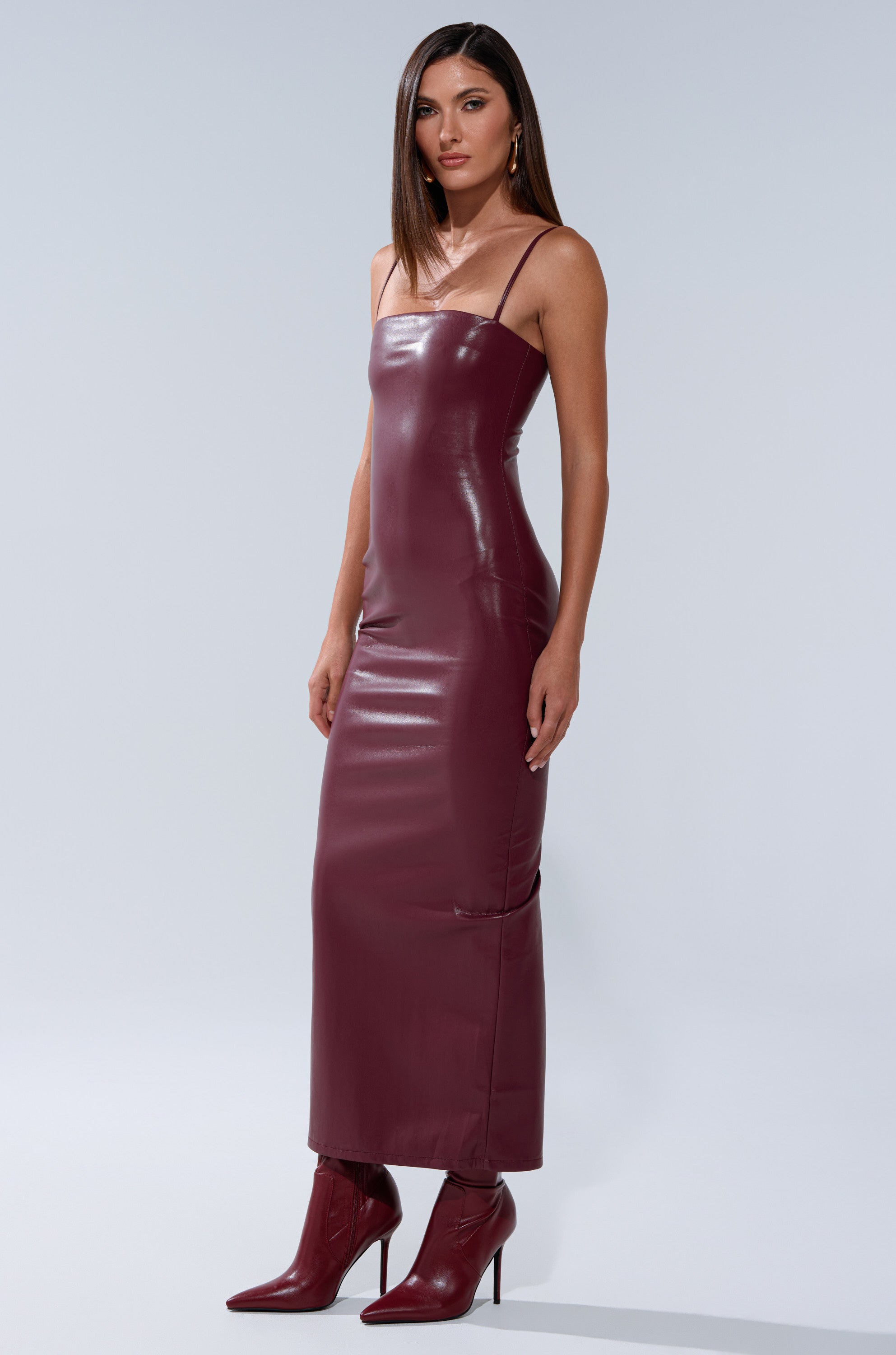 A woman models the SHAI PU MIDI DRESS IN BURGUNDY, a sleeveless, fitted faux leather dress with thin straps, styled with matching pointed-toe high-heeled boots against a plain light gray background. 4