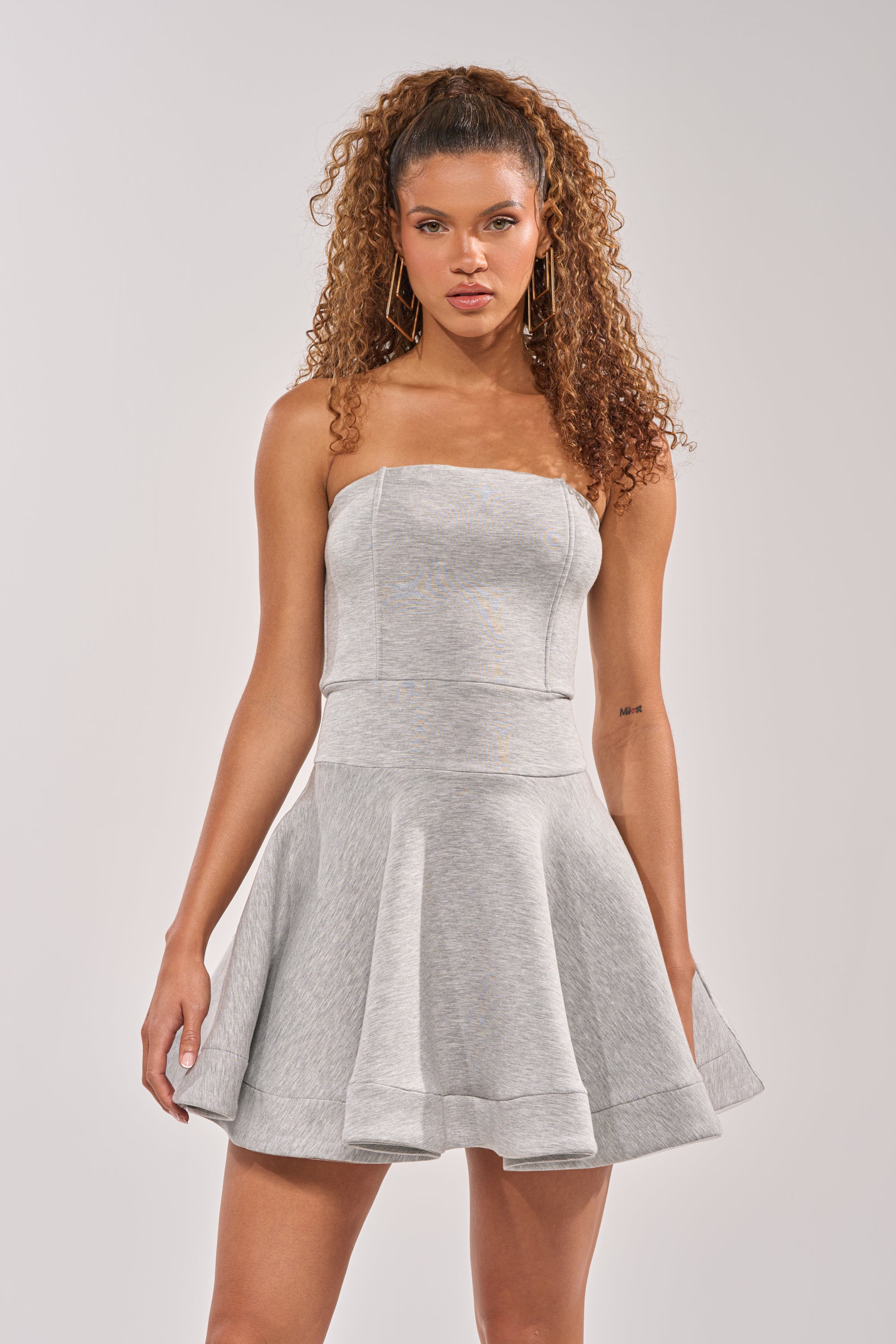 A woman with curly hair wears the SWEET NOTHINGS STRAPLESS MINI DRESS in light grey, standing facing forward against a plain, light background. 1
