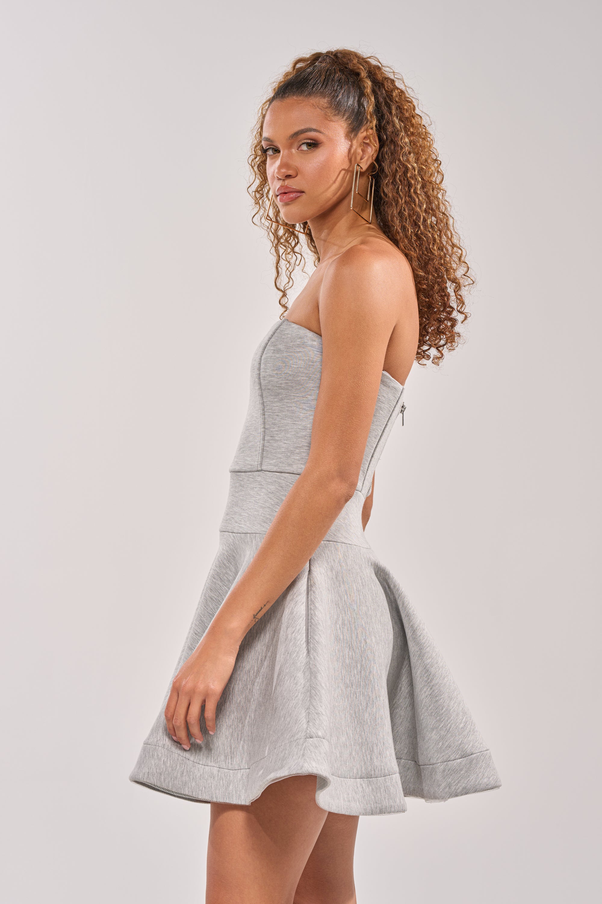 A woman with curly hair wears the SWEET NOTHINGS STRAPLESS MINI DRESS in light gray, featuring a fitted bodice and flared skirt, as she stands sideways looking over her shoulder against a plain light background. 2
