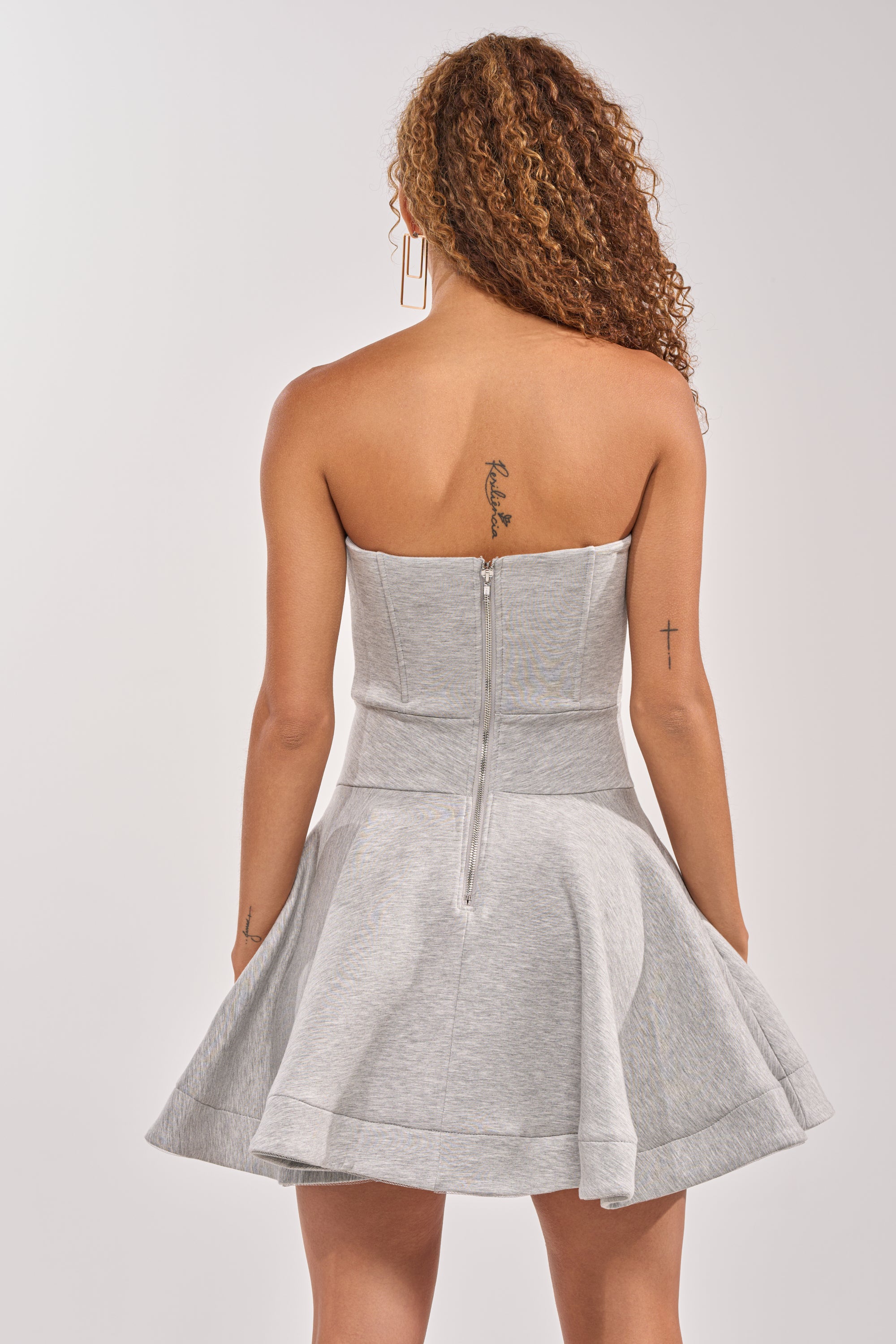 A woman with curly hair, tattoos on her right shoulder and left forearm, is seen from behind wearing the SWEET NOTHINGS STRAPLESS MINI DRESS—a heather gray, flared skater mini dress featuring two visible zippers. 3