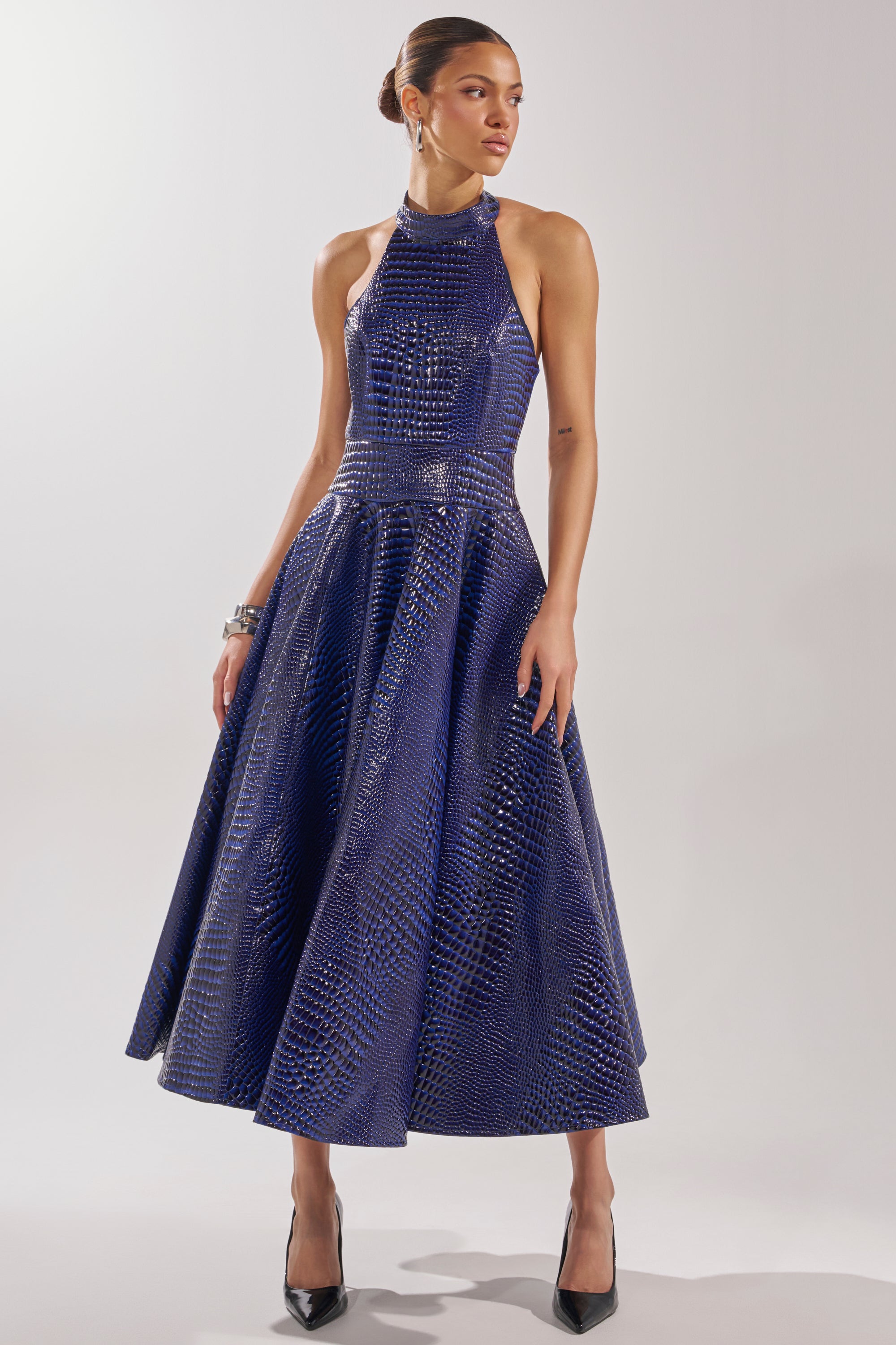 A woman models the VEE FAUX LEATHER CROC MIDI DRESS, a sleeveless, shiny dark blue crocodile-embossed piece, paired with black high heels. She poses gracefully with one hand on her skirt against a plain background.