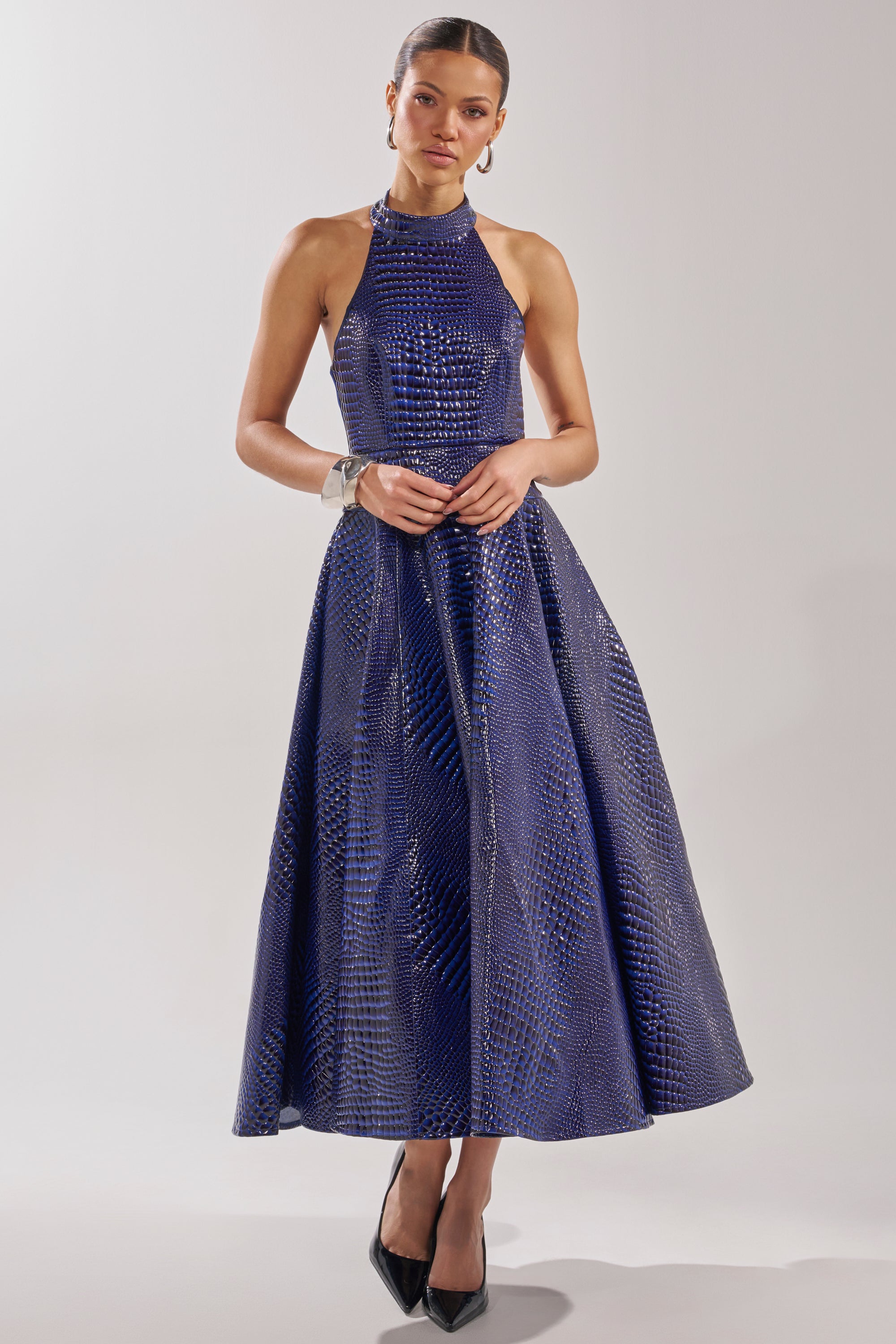 A woman stands confidently in black heels, wearing the VEE FAUX LEATHER CROC MIDI DRESS—a sleeveless royal blue dress with a subtle textured pattern—against a plain light background.