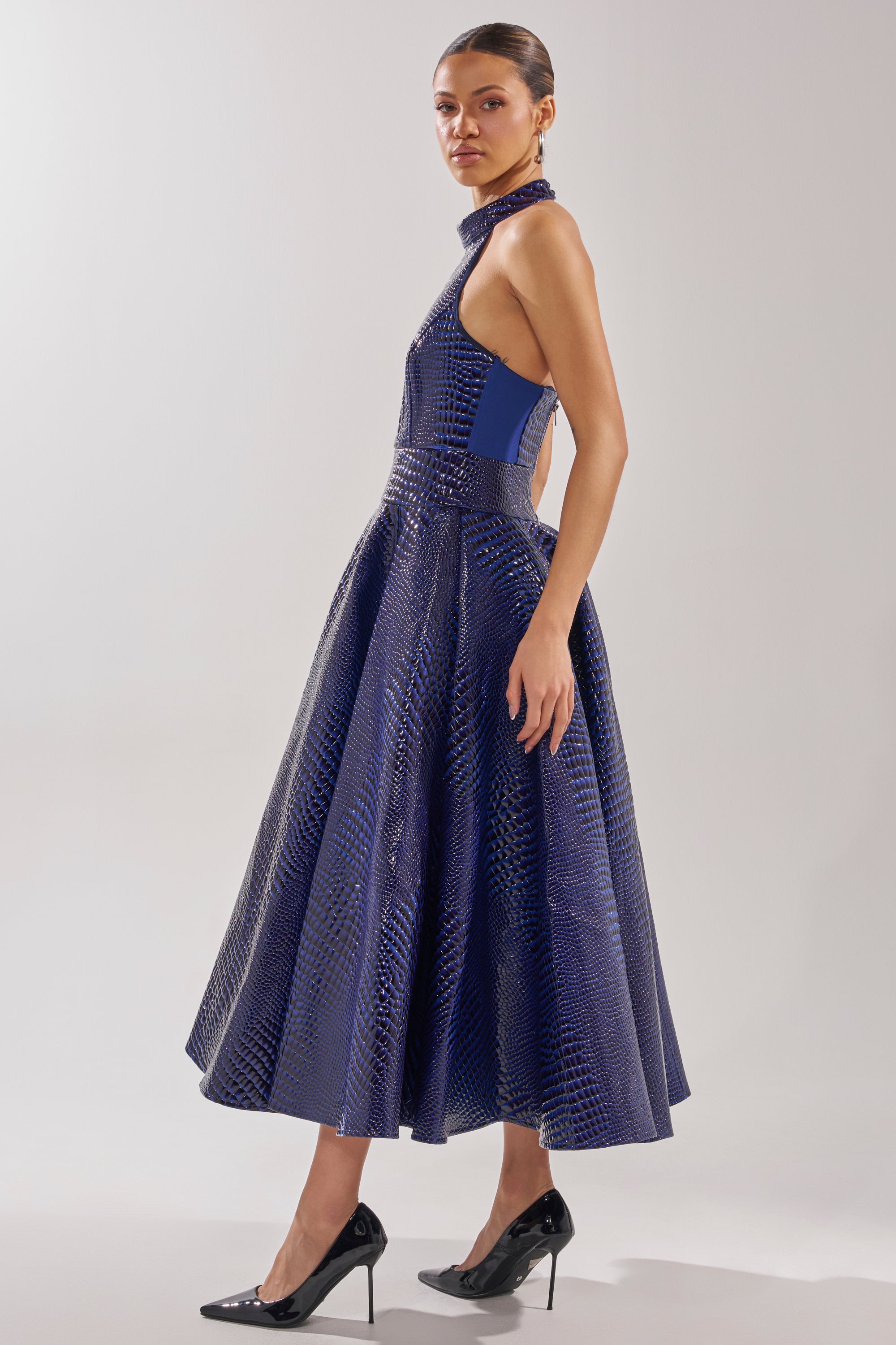 A woman in profile wears the VEE FAUX LEATHER CROC MIDI DRESS, a sleeveless, royal blue, ankle-length dress with a flared skirt, open back, and crocodile-textured pattern. She pairs it with high heels against a plain light background.