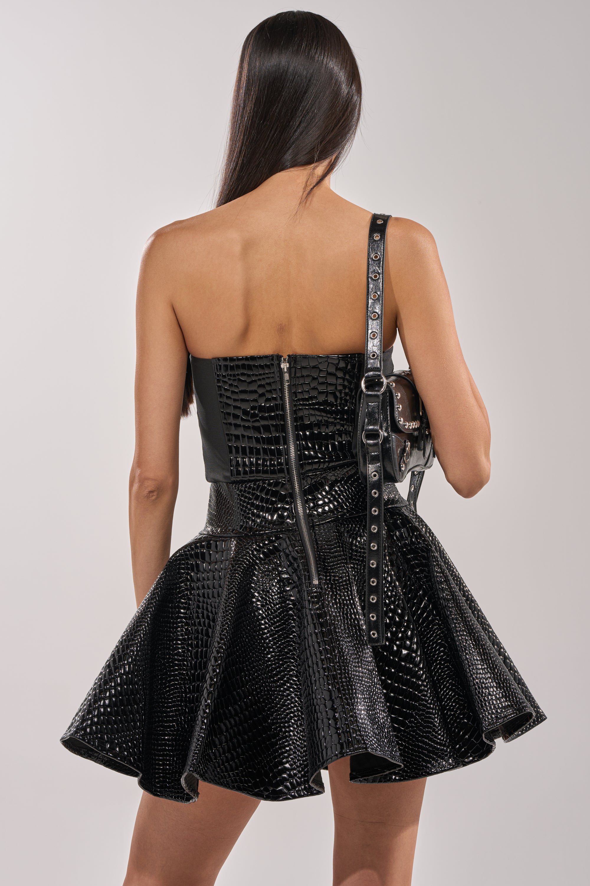 A woman with long straight hair, seen from behind, wears the VAL Faux Leather Mini Dress in Croc—strapless and black with a flared skirt and front zipper—and carries a black studded handbag over her shoulder. 3